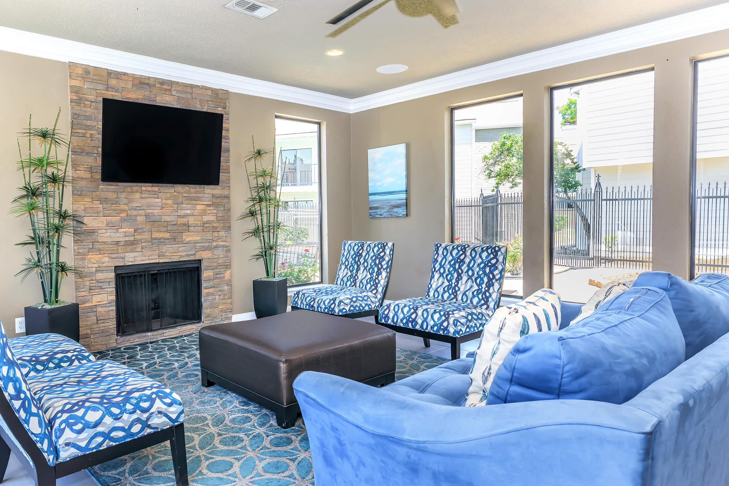 A bright living room with a cozy arrangement featuring a blue sofa, patterned chairs, and a dark coffee table. There's a stone fireplace with a TV mounted above it, and large windows letting in natural light. Decorative plants are placed in each corner, enhancing the modern aesthetic.