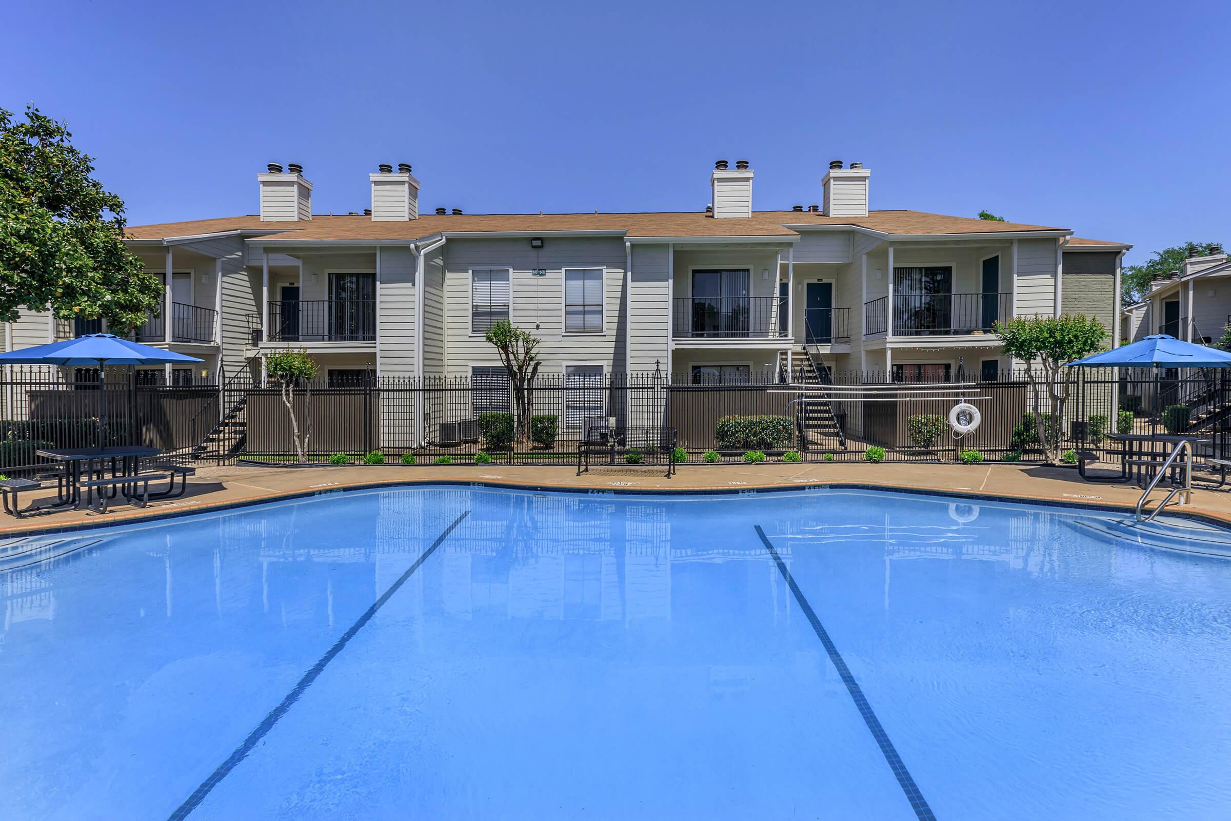 A clear blue swimming pool in the foreground, with lounge chairs and umbrellas around it. In the background, there are two-story apartment buildings with a light exterior. The setting is sunny, with trees and landscaped greenery visible. The scene conveys a relaxing, residential atmosphere.