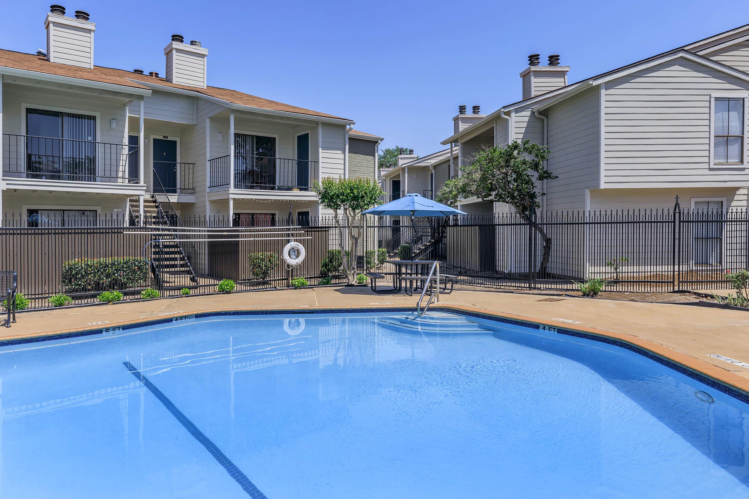 A serene outdoor space featuring a clear blue swimming pool surrounded by a fenced area. In the background, there are two multi-story residential buildings with balconies, and a shaded seating area under an umbrella. Lush greenery and well-maintained landscaping enhance the setting.