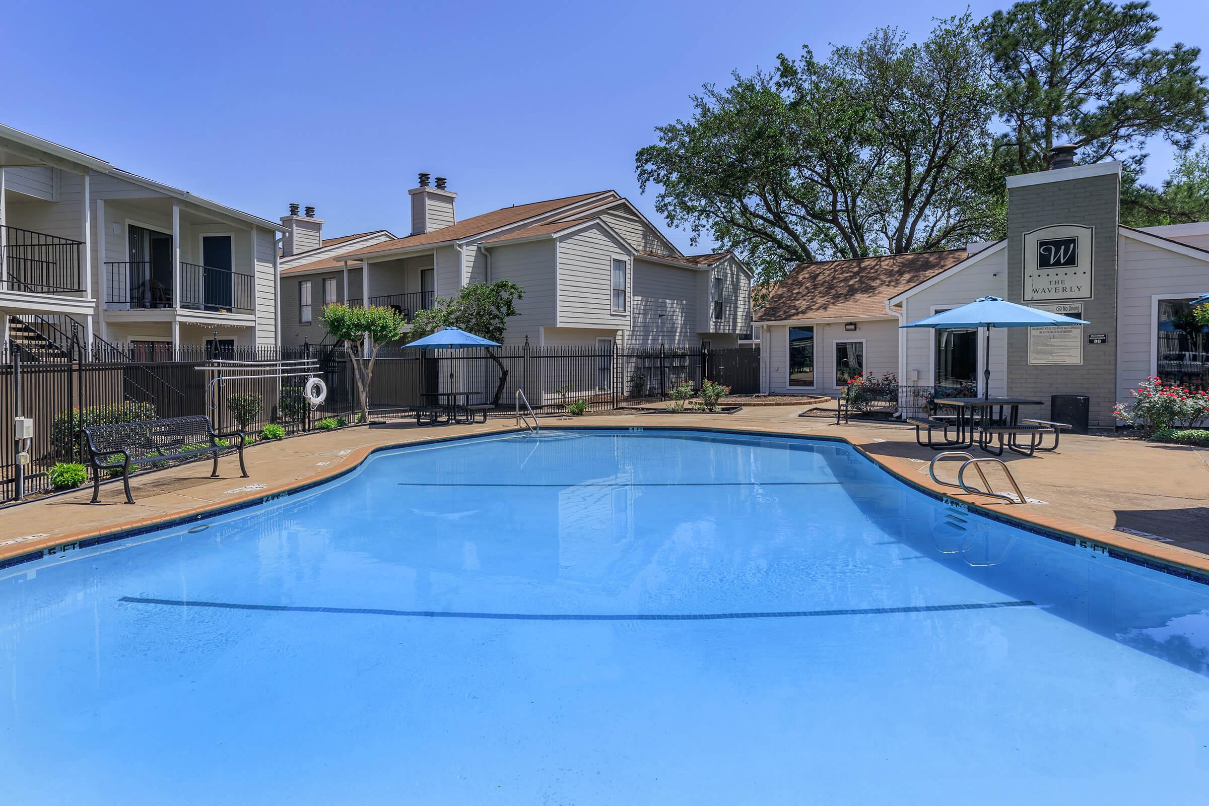 A clear, blue swimming pool surrounded by a patio area, with lounge chairs and shade umbrellas. Residential buildings with balconies can be seen in the background, along with well-maintained landscaping. The scene is bright and inviting, suggesting a relaxing outdoor space.
