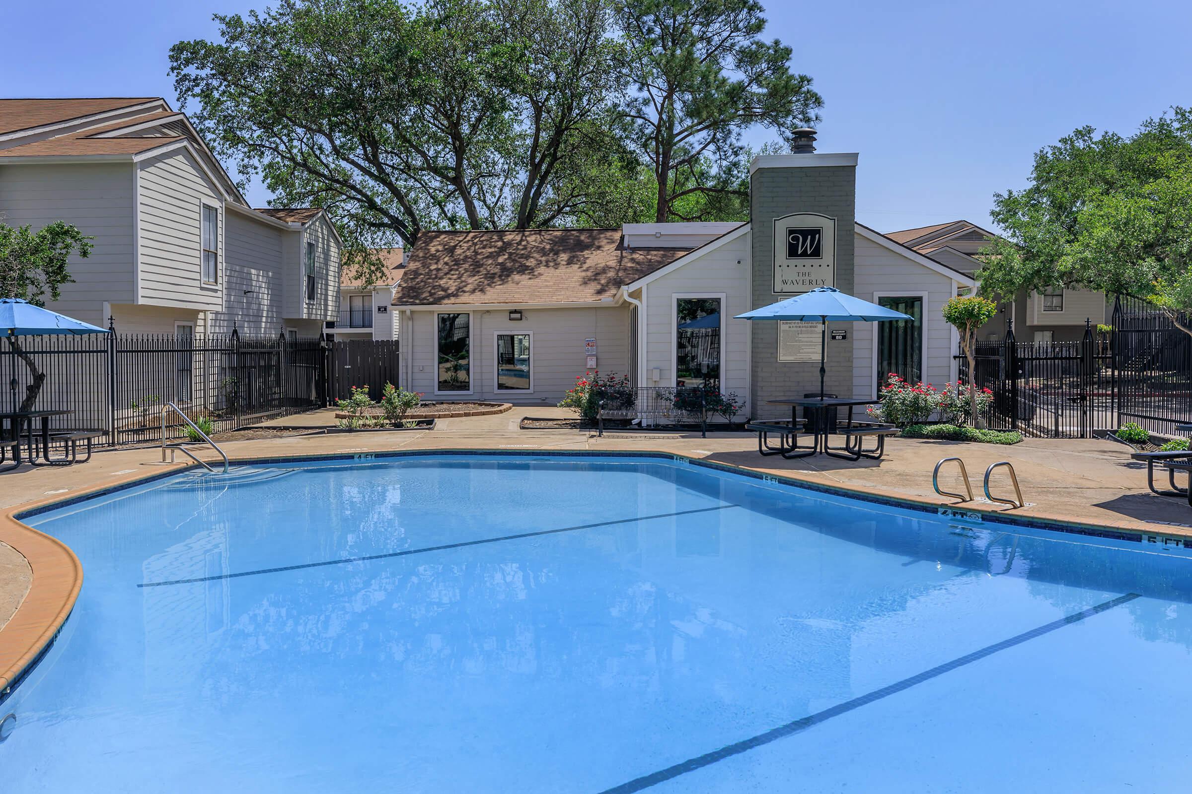 A bright swimming pool with clear water, surrounded by a concrete deck and lounge areas. In the background, there’s a single-story building with a sign, alongside trees and umbrellas providing shade. The scene conveys a relaxed, inviting atmosphere ideal for leisure and recreation.