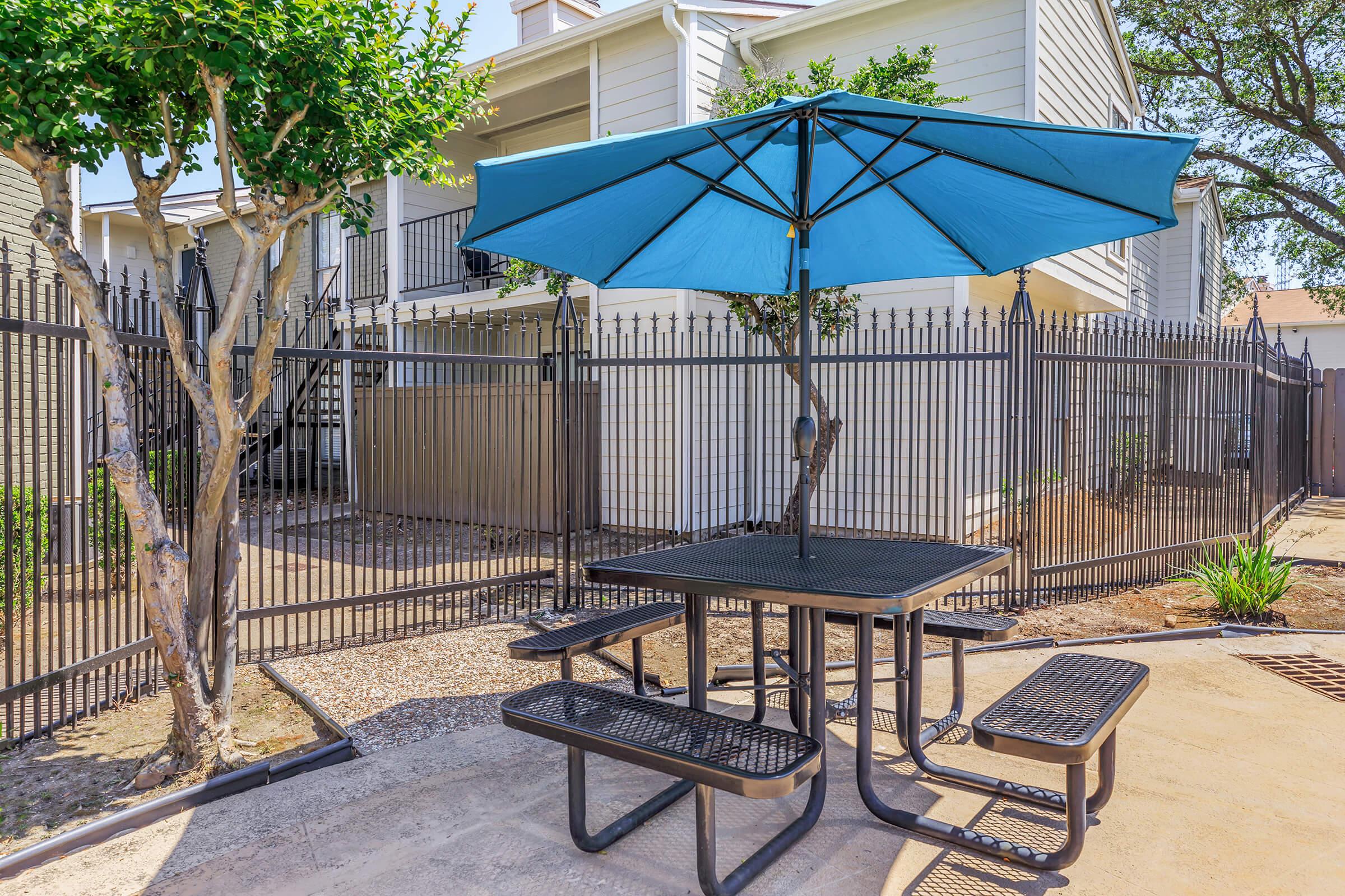 A shaded outdoor seating area featuring a black metal picnic table with attached benches, under a large blue umbrella. The setting includes landscaped greenery and a fenced area with apartment buildings in the background. The scene is bright and sunny, ideal for relaxation or social gatherings.