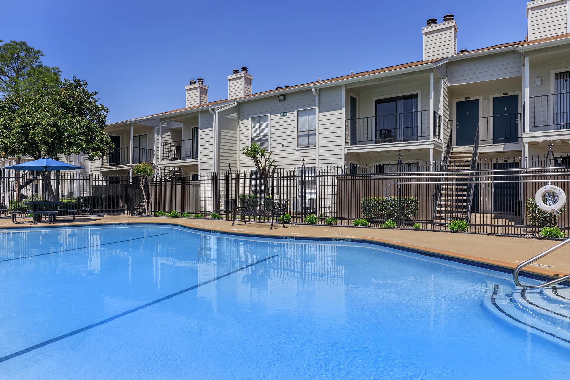 A clear blue swimming pool reflects the sky, surrounded by a fenced patio area. In the background, there are two-story apartment buildings with balconies. Lush greenery and trees add to the inviting atmosphere, while patio furniture is arranged nearby for relaxation.