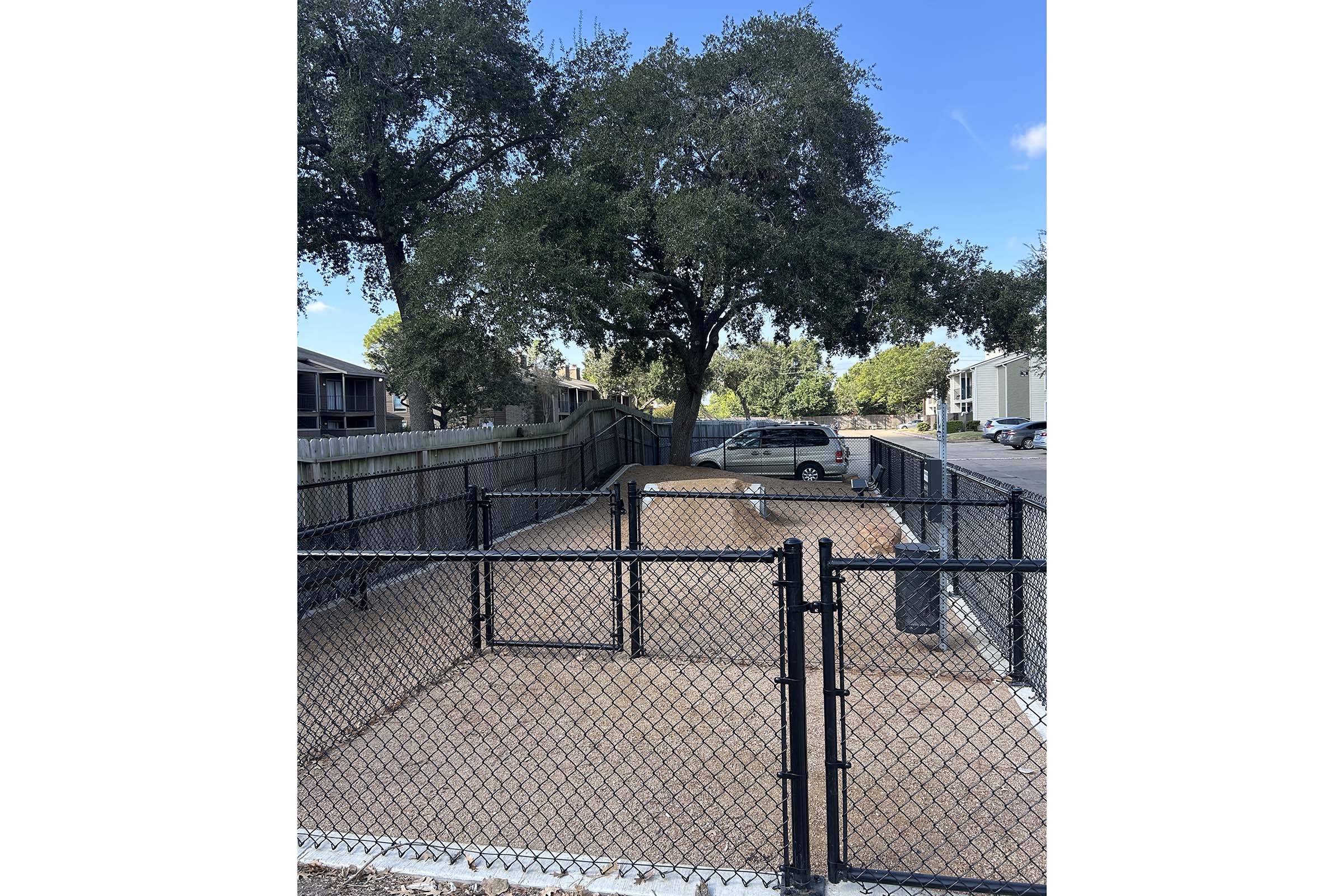 A fenced outdoor area with a sandy surface, partially shaded by a large tree. The space is surrounded by a black chain-link fence and features a few scattered rocks. Nearby, there is a parked vehicle and residential buildings in the background under a clear blue sky.
