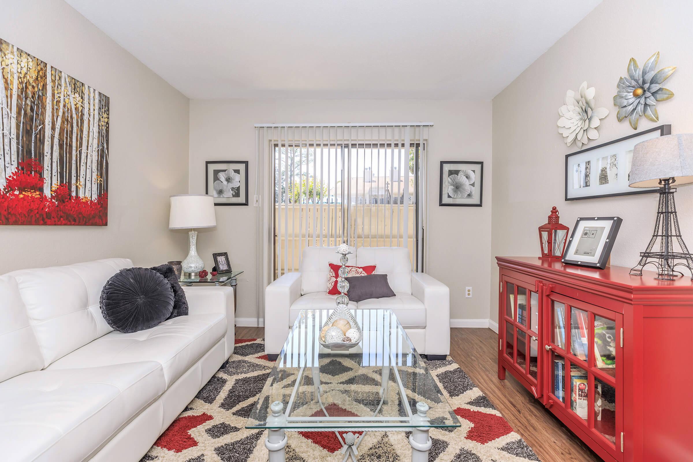 A cozy living room featuring a white leather sofa and armchair. A glass coffee table sits on a patterned rug, while decorative elements include a red cabinet, lamp, and wall art with trees and flowers. Natural light filters through vertical blinds, creating a warm atmosphere.