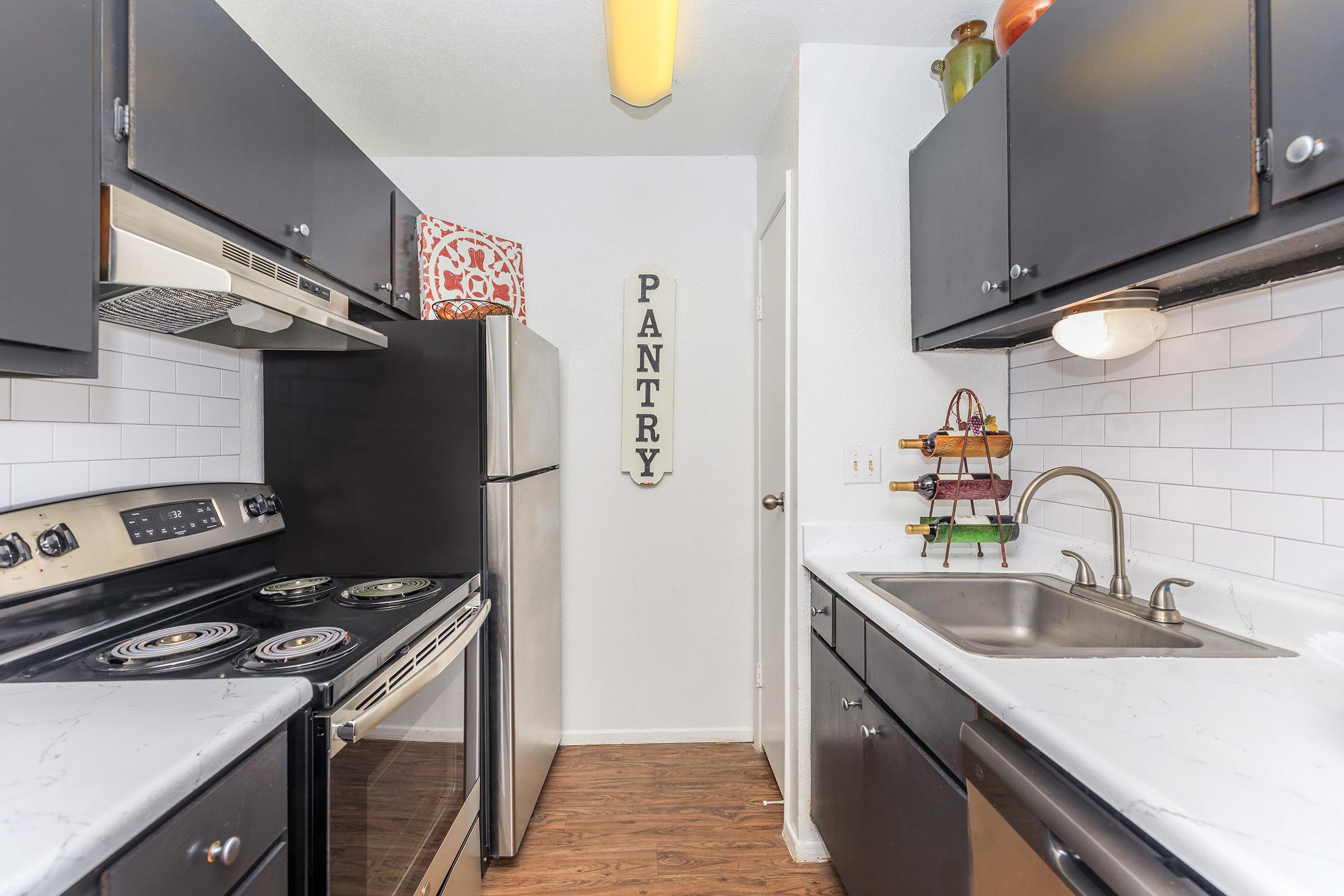 A modern kitchen featuring dark cabinetry, stainless steel appliances including a refrigerator and stovetop, and a white tiled backsplash. The kitchen has a functional layout with a sink, countertop, and a pantry sign on the wall, along with decorative items on the shelves.