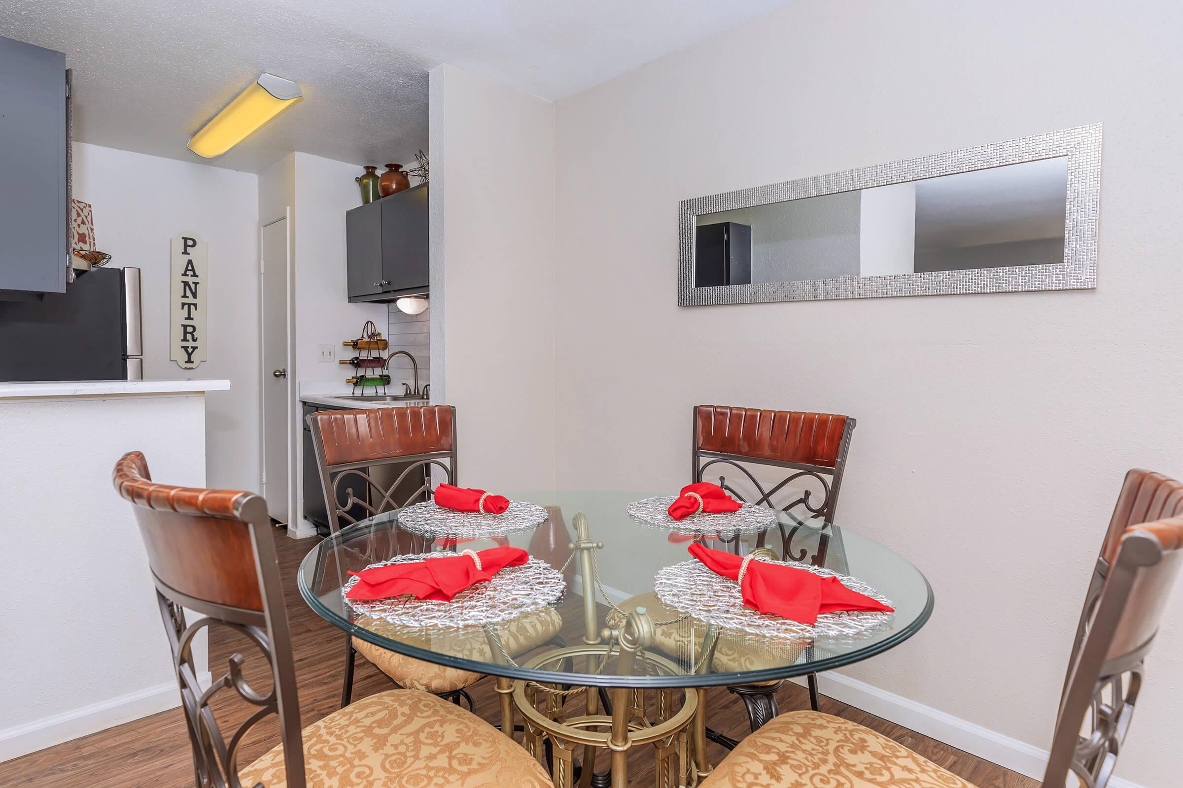 A dining area featuring a round glass table set with red napkins, surrounded by metal chairs with decorative cushions. In the background, there's a kitchen area with a pantry door, shelves, and modern cabinets. A mirror on the wall adds depth to the space. The décor is simple and elegant.