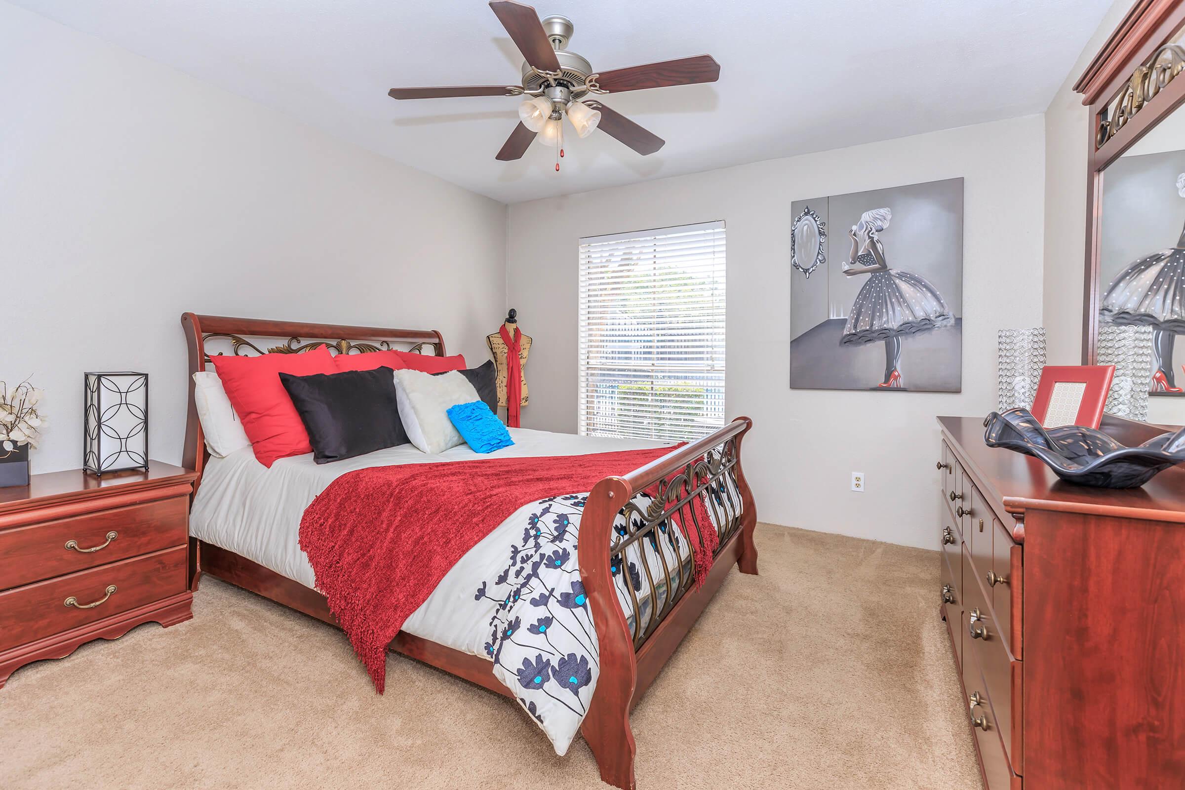 A cozy bedroom featuring a wooden bed with a red and blue decorative arrangement, fluffy bedding, and two nightstands. A dresser with a mirror is present, along with a wall art piece of a dress form and a framed photo. The room has natural light coming from a window with blinds.
