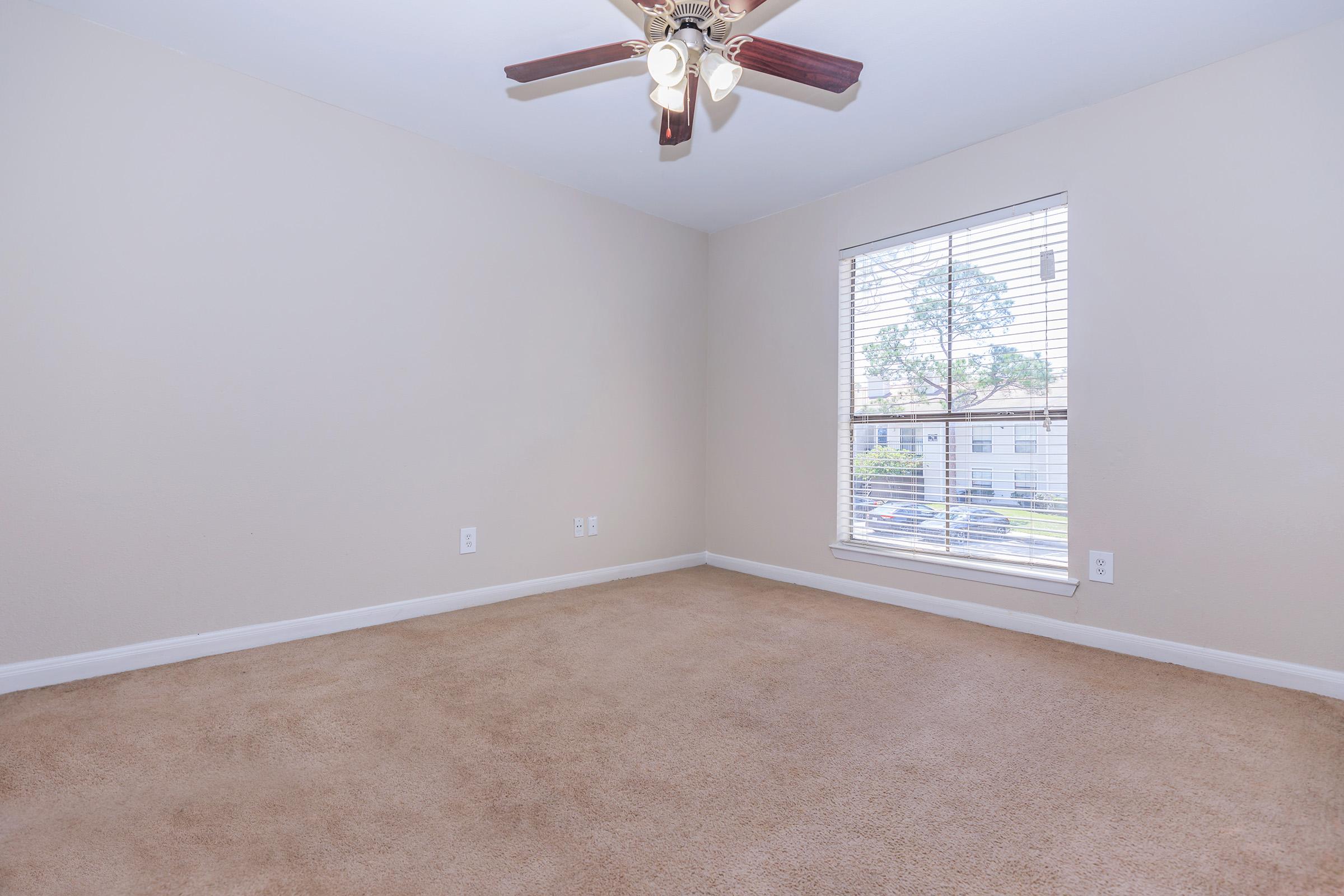 Empty room featuring beige walls and carpet, a ceiling fan with wooden blades, and a window with blinds allowing natural light to enter. The window overlooks an outside area, creating a bright and open atmosphere in the space.