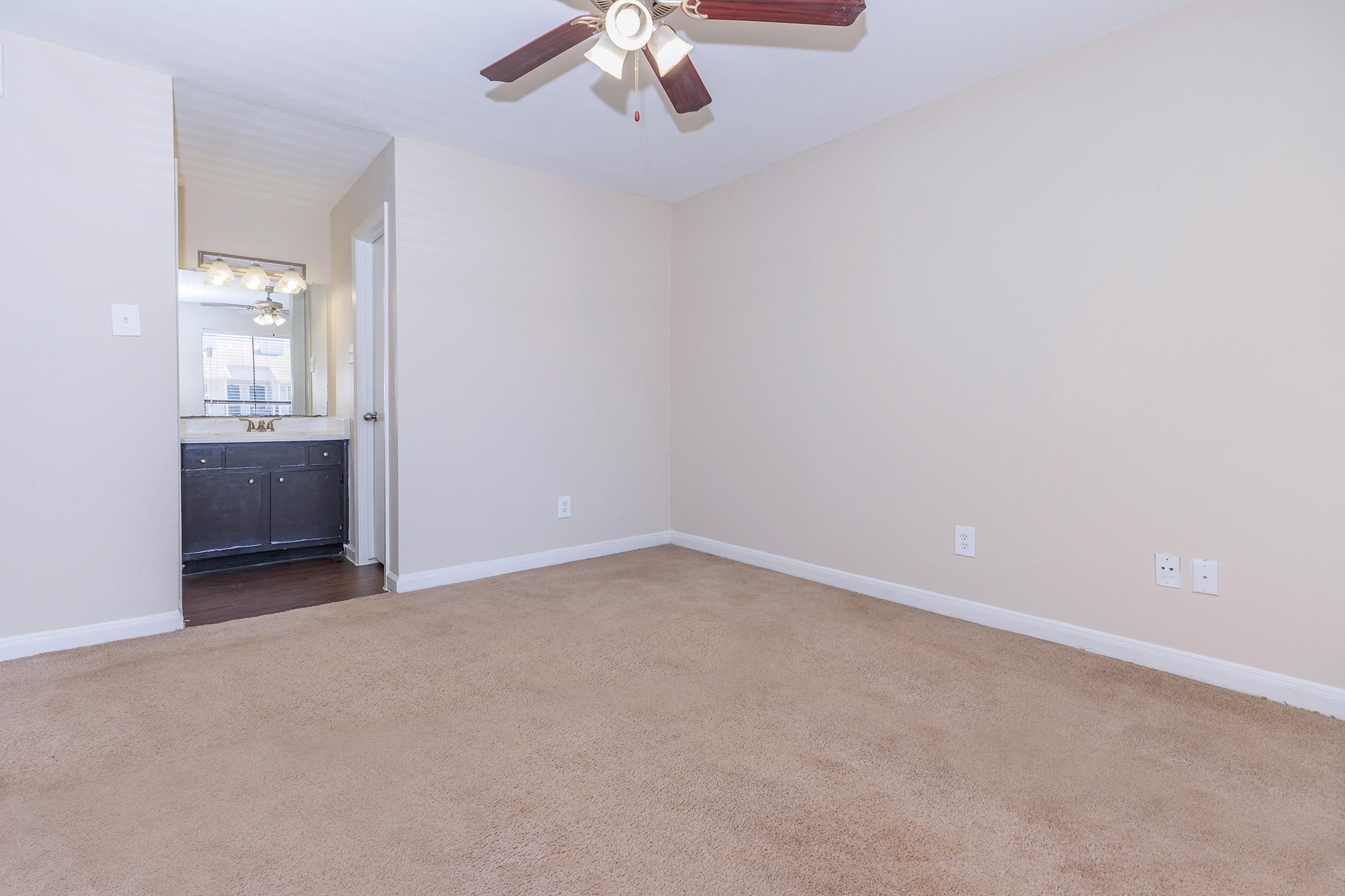 A spacious, empty room with light beige walls and carpet. A ceiling fan is visible, and there's a doorway leading to a small bathroom area with a mirror and dark cabinetry. Natural light is coming from the doorway, creating a bright atmosphere.