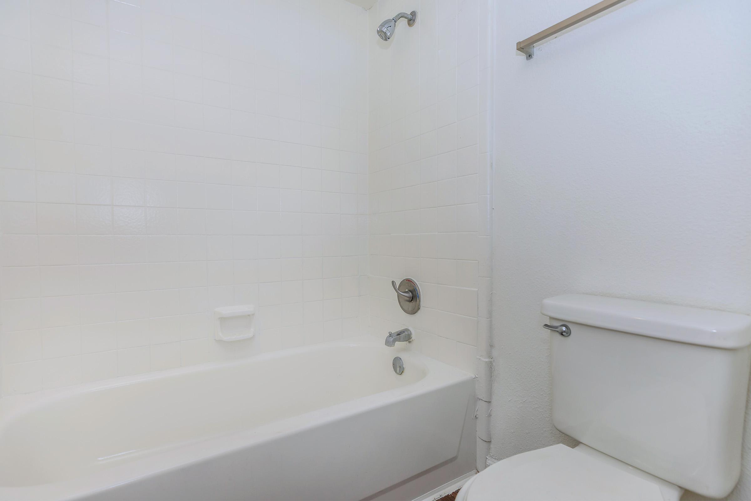 A clean, simple bathroom scene featuring a white bathtub with a faucet and a showerhead, along with a white toilet. The walls are tiled in white, creating a bright and spacious appearance. The overall design is minimalist and modern, with no additional decorations or clutter present.