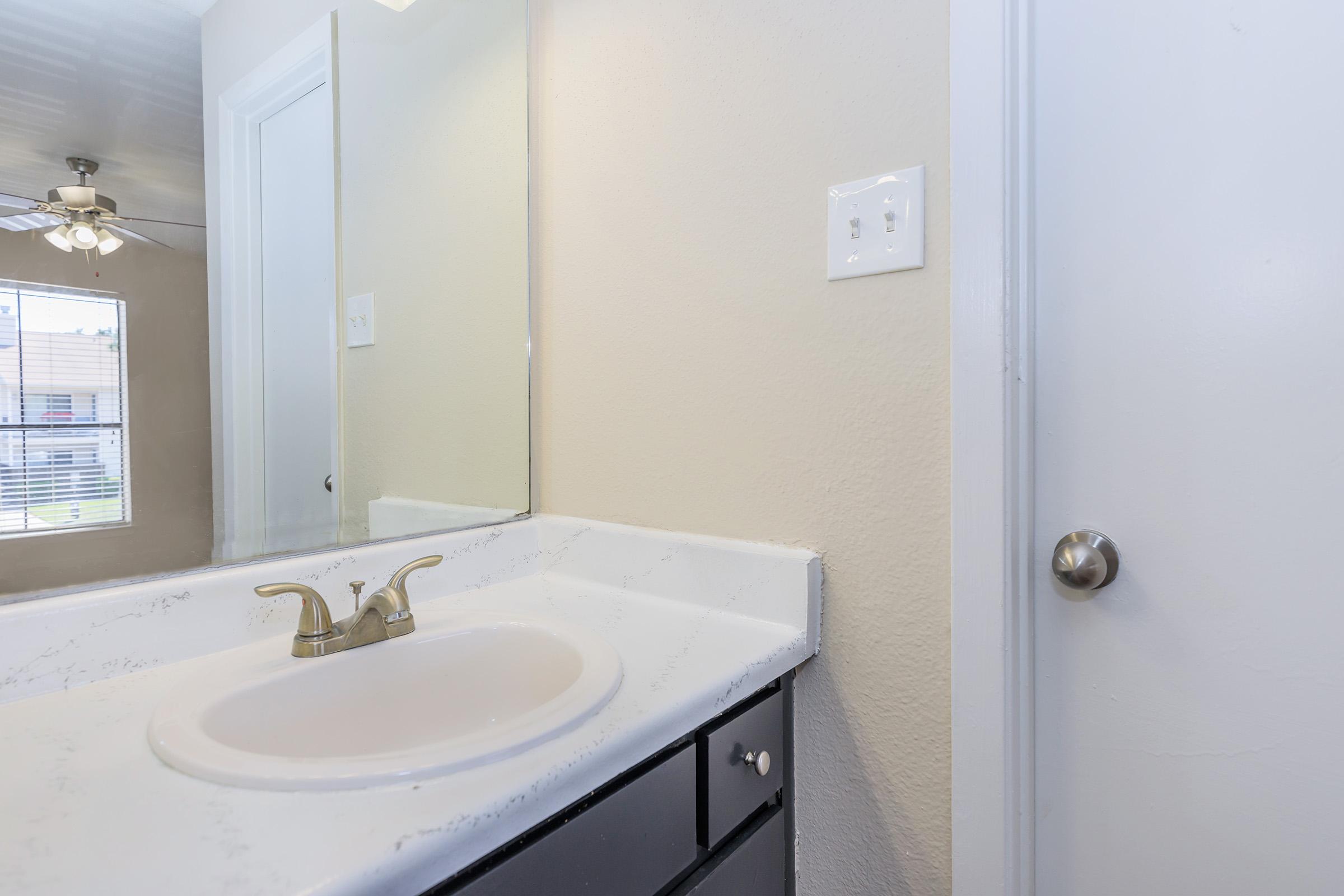 A bathroom sink with a simple faucet and countertop, positioned near a mirror. The wall is a light neutral color, and there is a door visible on the right side of the image. Natural light streams in from a window on the left, adding brightness to the space. A ceiling fan is visible in the background.
