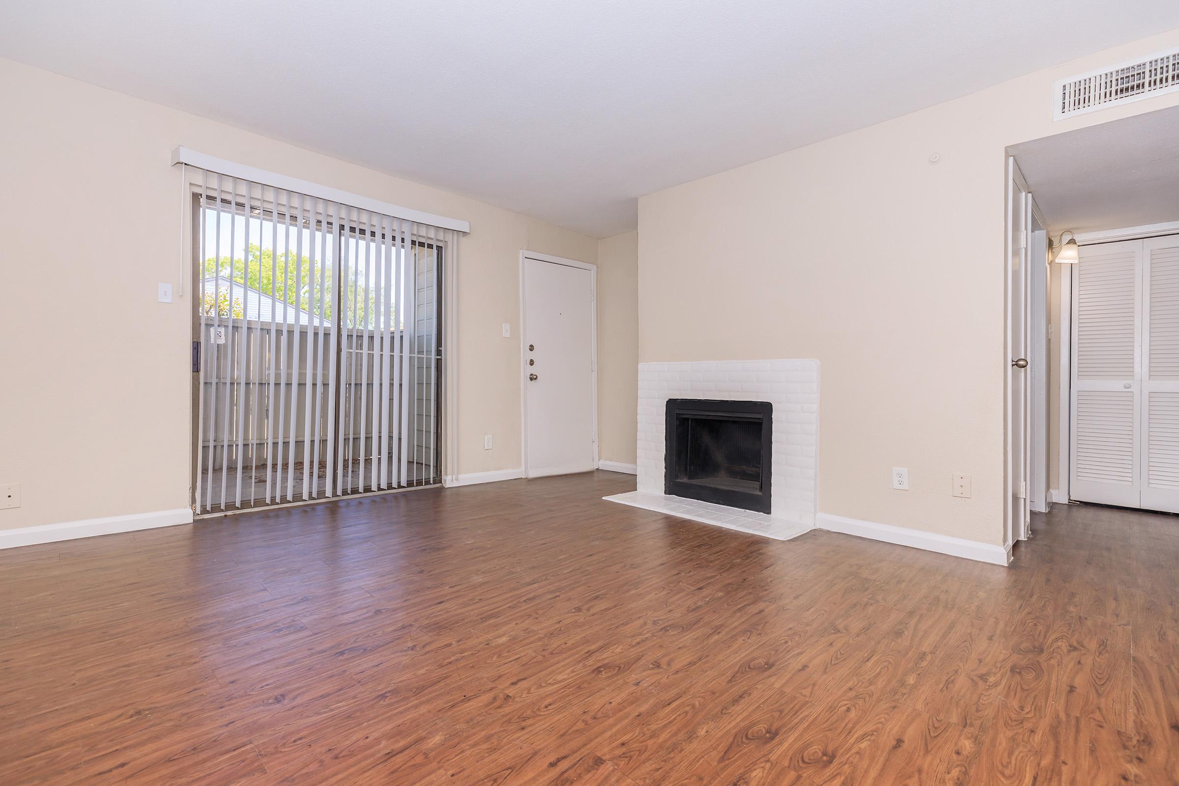 Spacious living room with wood-style flooring, a white brick fireplace, and a sliding glass door leading to a balcony. Light-colored walls and natural light create a welcoming atmosphere. A closet is visible on the right, enhancing storage options.