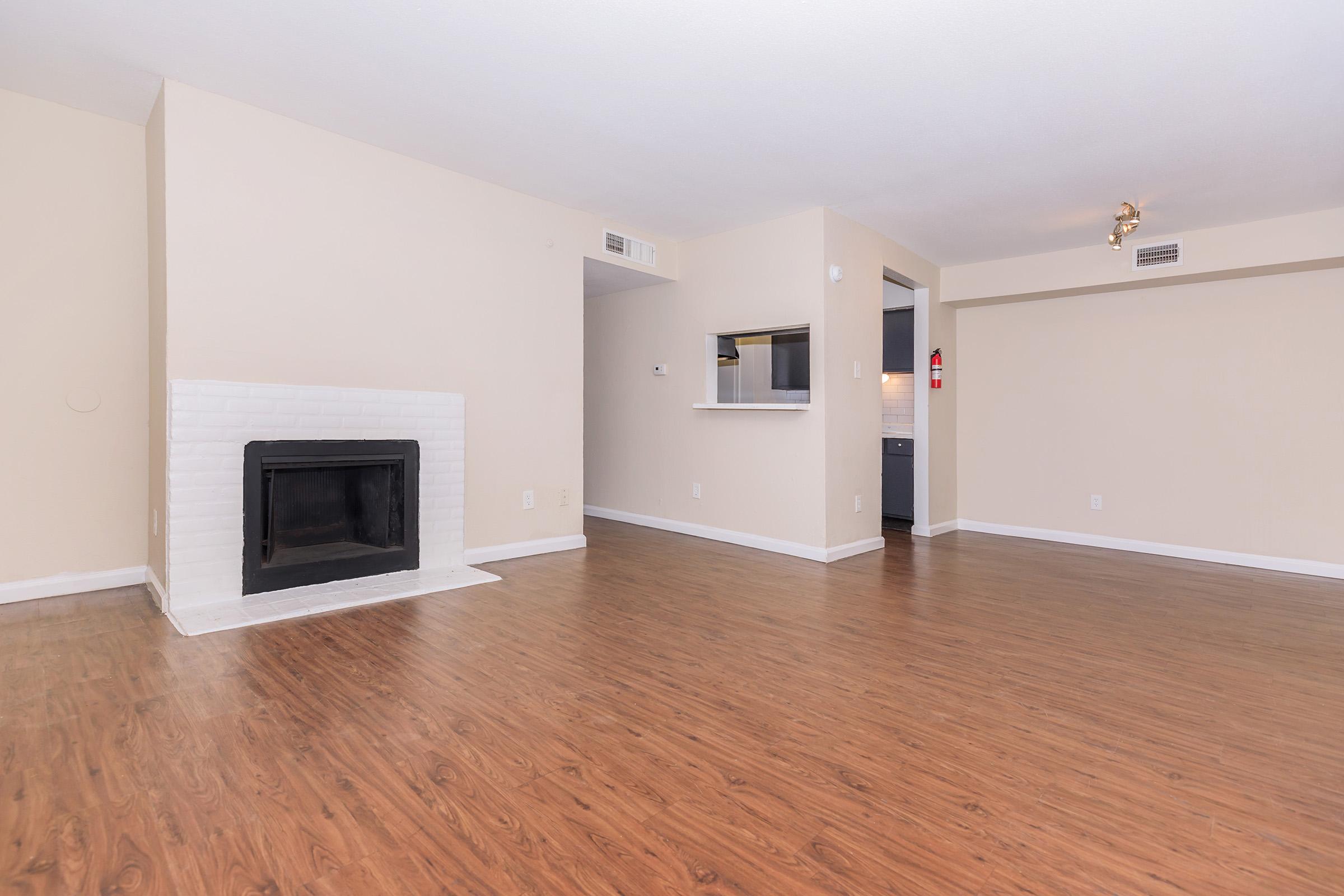 Spacious living area featuring a white brick fireplace, light beige walls, and light wood flooring. In the background, a small bar area opens into a modern kitchen, with a doorway leading to an unseen space. The room is well-lit with natural light and has a minimalist design.