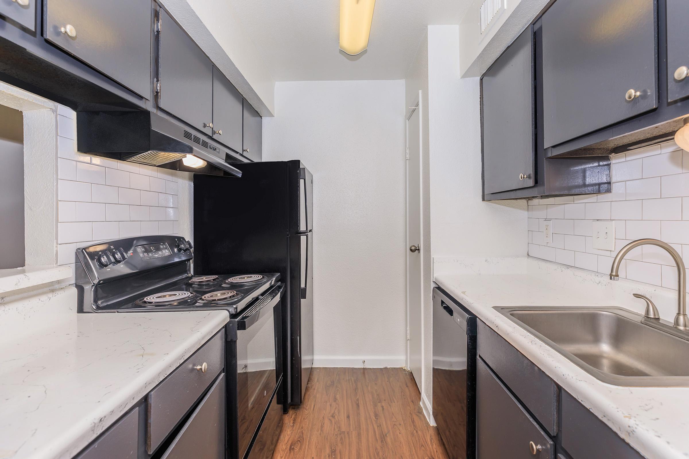 Modern kitchen featuring black cabinets, a black refrigerator, and an oven with a stovetop. The countertops are light-colored with a subtle pattern, and the walls are white with a tiled backsplash. A sink is positioned next to a window, and the floor is finished with wood-like laminate.