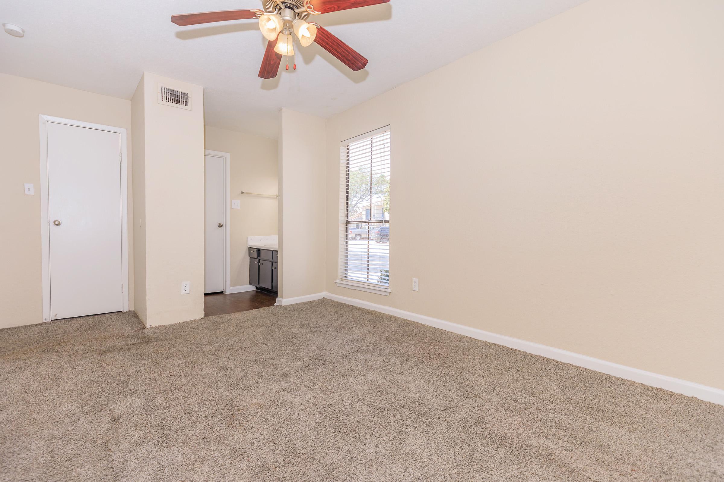 A clean, empty room with tan walls and carpet. It features a ceiling fan with four light bulbs, a window with blinds allowing natural light, and a doorway leading to another room. There is a small kitchenette area visible to the left.