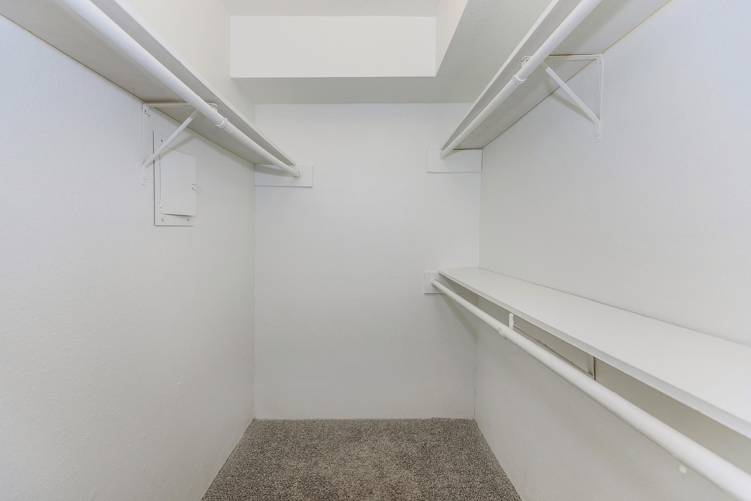 Empty closet featuring white walls, a single shelf, and two hanging rods on either side. The floor is covered with a light carpet, creating a minimalistic and uncluttered appearance.
