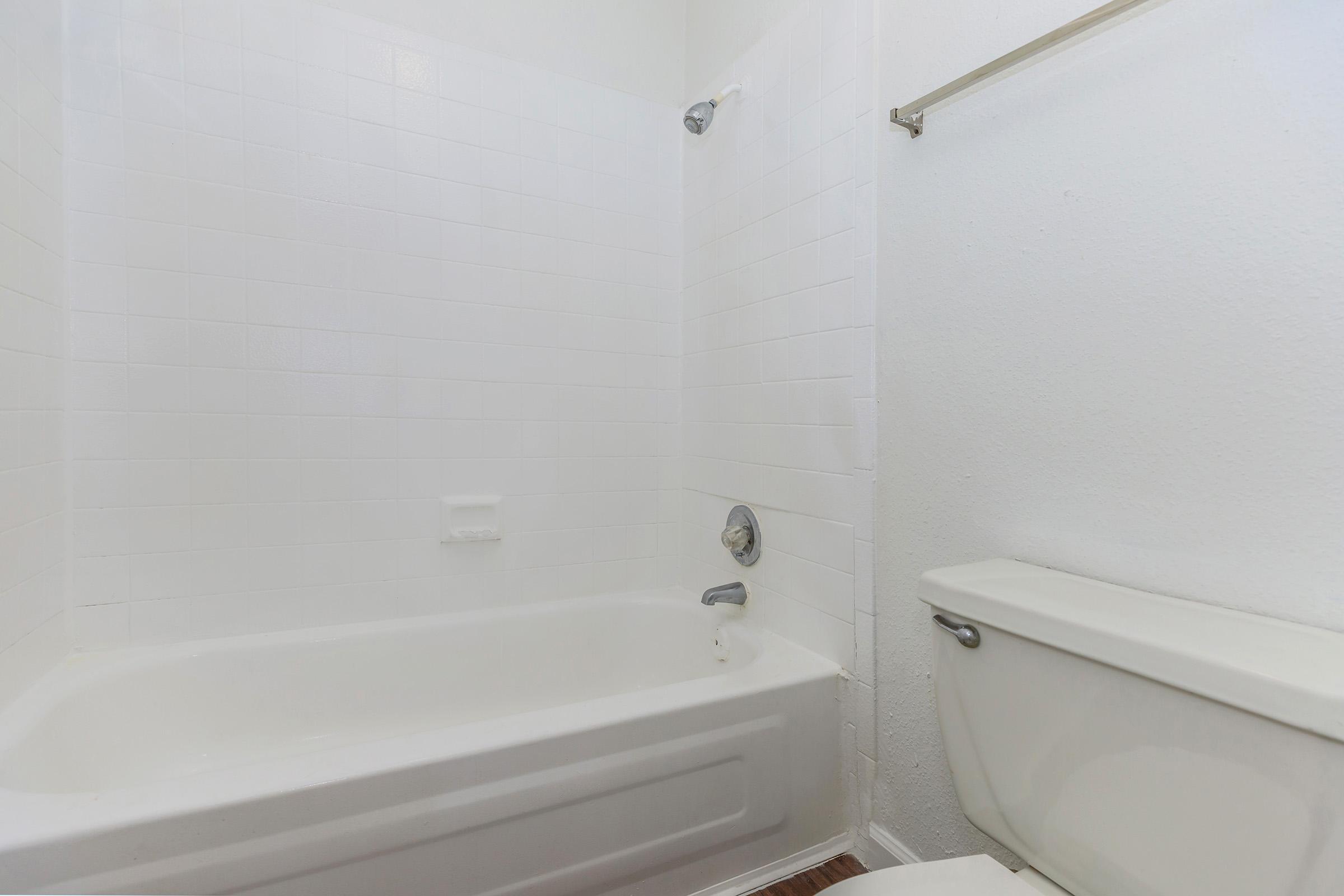 A clean, minimalistic bathroom featuring a white bathtub with a showerhead mounted above, set against plain white tiled walls. A toilet is positioned nearby, completing the simple, functional design of the space. The floor is wooden, adding warmth to the overall look.