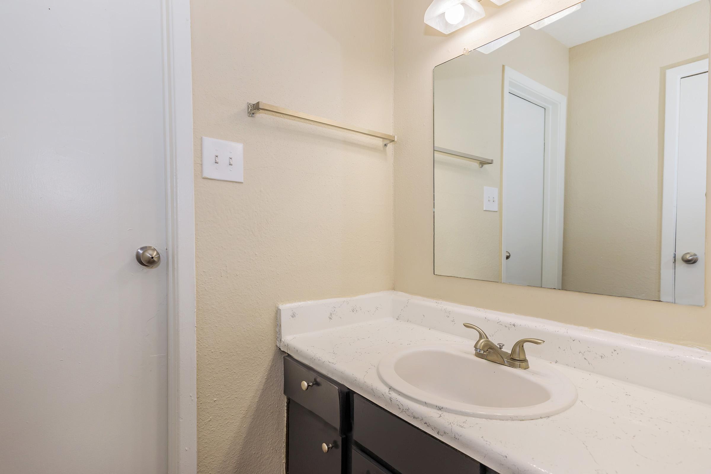 Bright bathroom featuring a light-colored wall, a large mirror above a white countertop with an integrated sink, and a brushed gold faucet. The cabinetry below the sink is dark-colored, and a towel bar is mounted on the wall. A door is visible to the left, indicating access to another room.