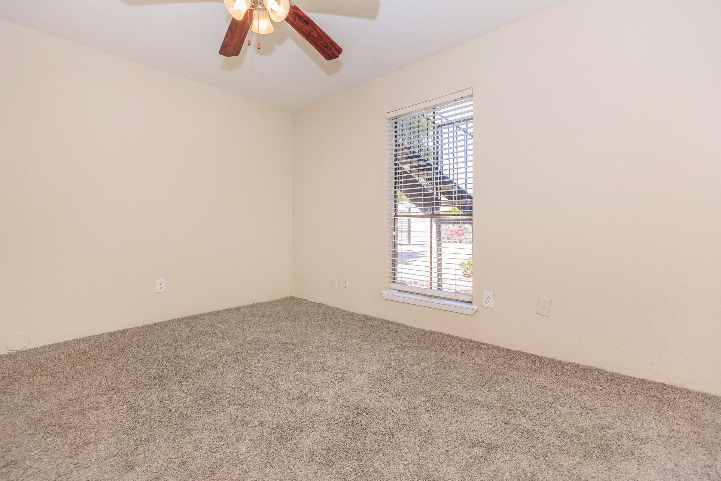 A vacant room featuring light beige walls and carpeted flooring. The room has a ceiling fan with wooden blades and a window with horizontal blinds, allowing natural light to enter. The corner of the room is empty, emphasizing the spacious feel.