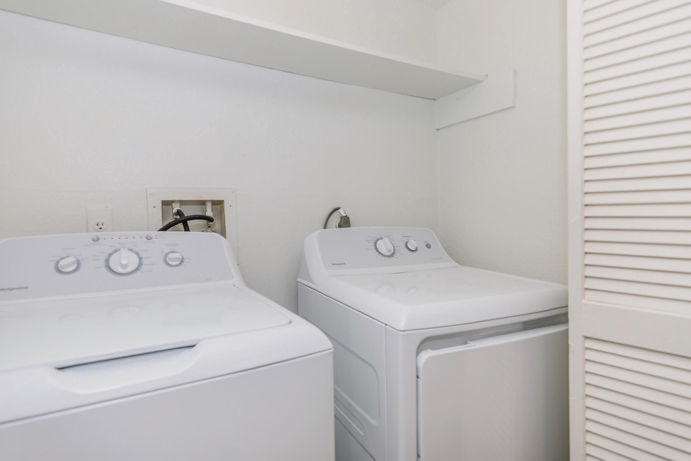 A clean laundry room featuring a front-loading washing machine and a stacked dryer, set against a plain white wall. The appliances are neatly arranged side by side, and the room has a bright, minimalist aesthetic with ample space around them.