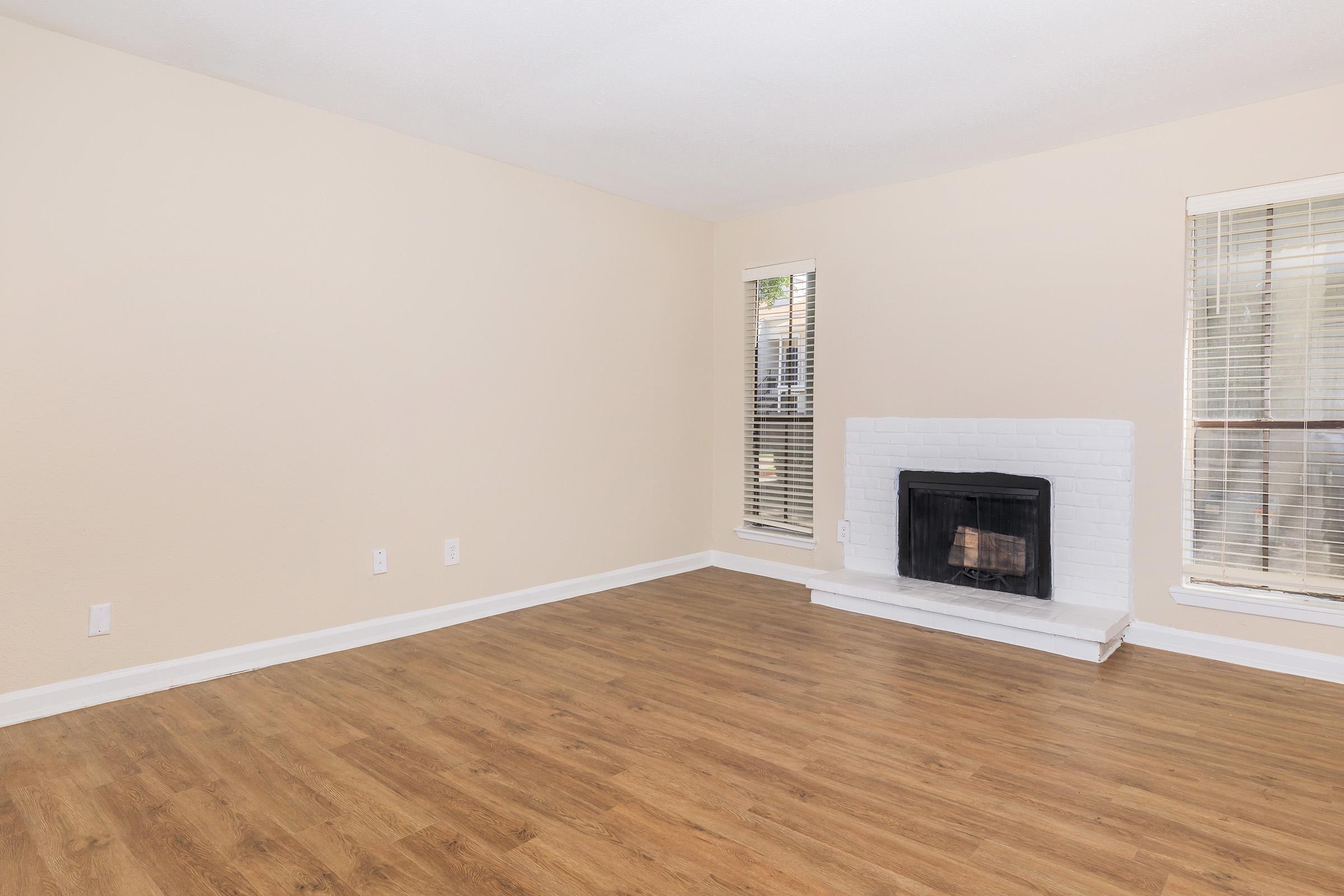 A spacious, empty living room featuring light-colored walls, warm wood-like flooring, and a white brick fireplace. Large windows with blinds allow natural light to fill the space, creating an inviting atmosphere suitable for decorating and personalization.