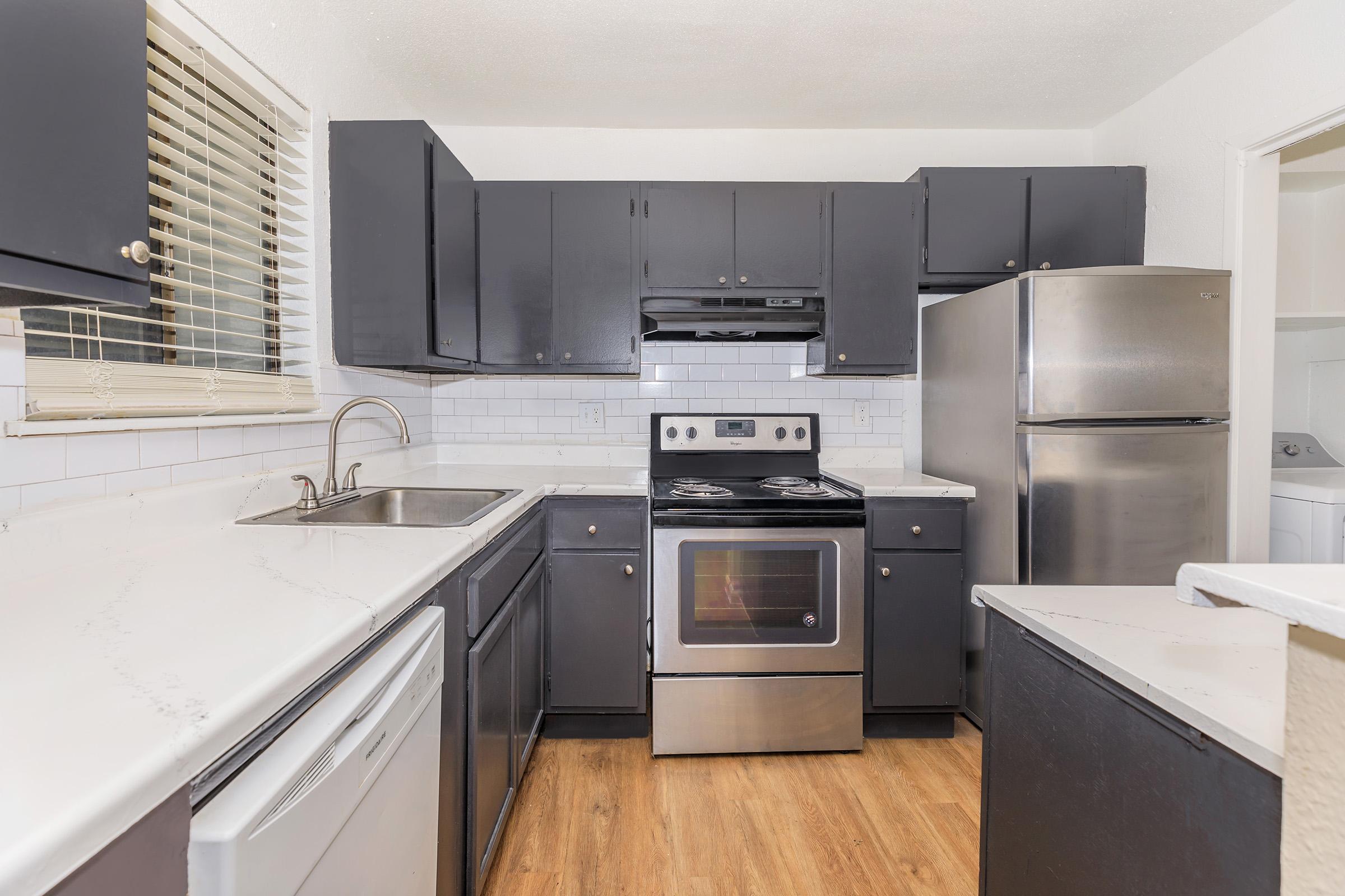 Modern kitchen featuring dark cabinetry, a stainless steel refrigerator, an electric stove with an oven, and a sink with a view of the exterior. The countertops are light-colored, and the flooring is wood-like. Large window above the sink provides natural light.