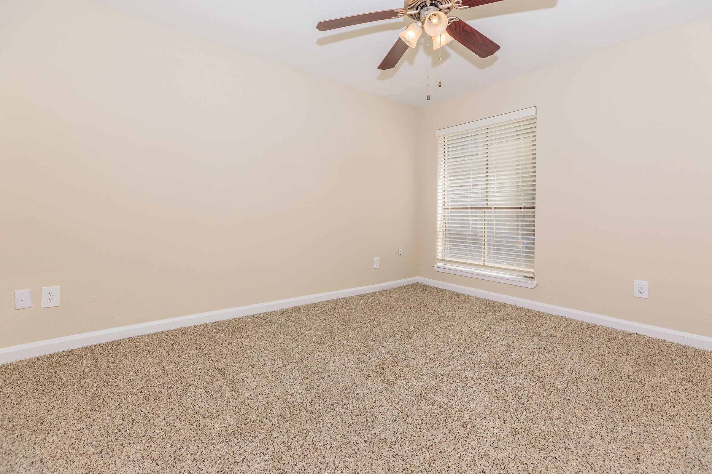 A sparsely furnished room with beige walls and light brown carpet. There is a ceiling fan with wooden blades and a window with blinds allowing natural light to enter. The overall ambiance is neutral and minimalistic, ideal for customization or redecorating.
