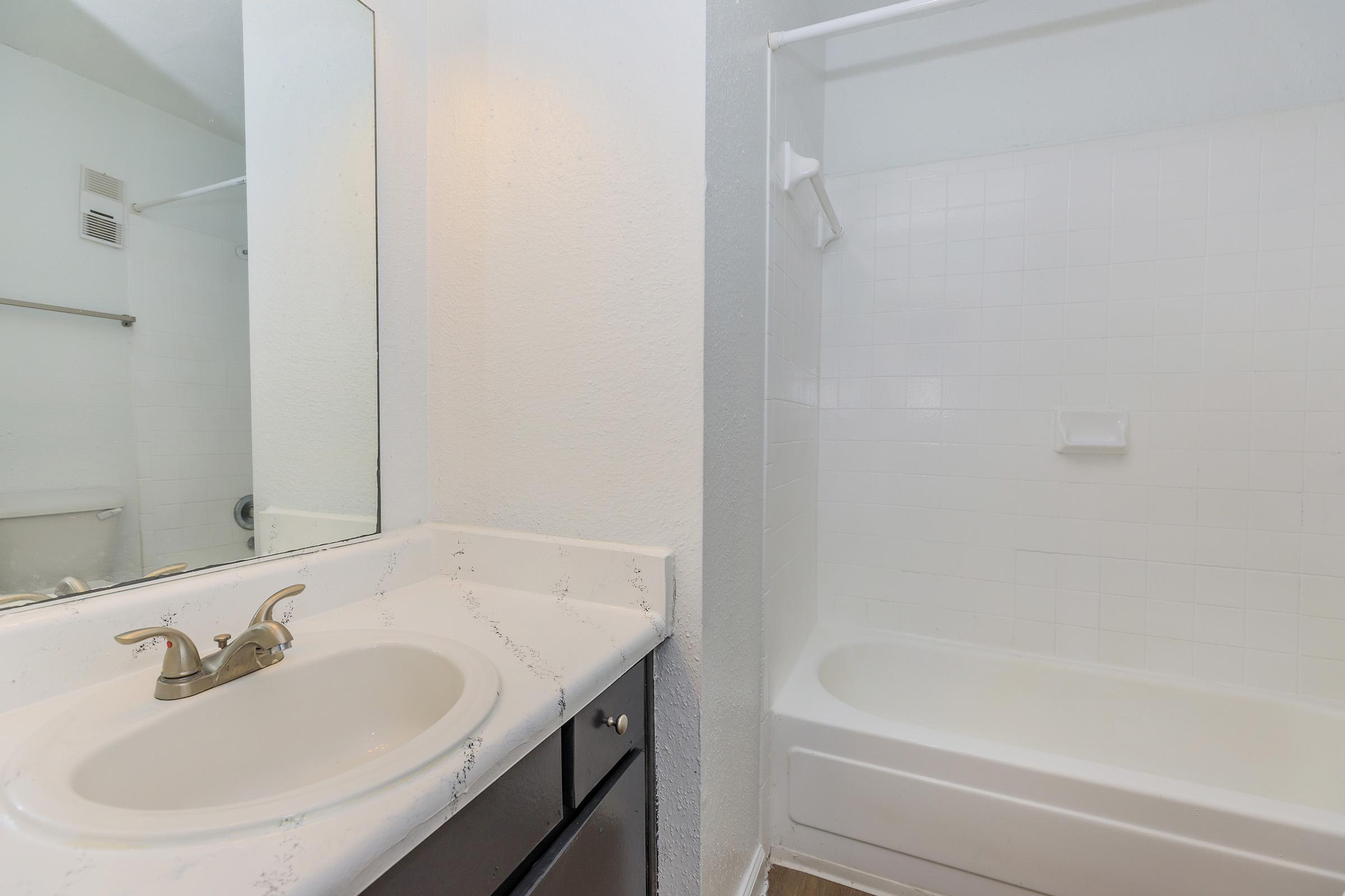 A clean bathroom featuring a white bathtub with a shower, a single sink with a marble countertop, and a mirror above the sink. The walls are painted light color, and the flooring is a neutral shade, creating a bright and fresh atmosphere.