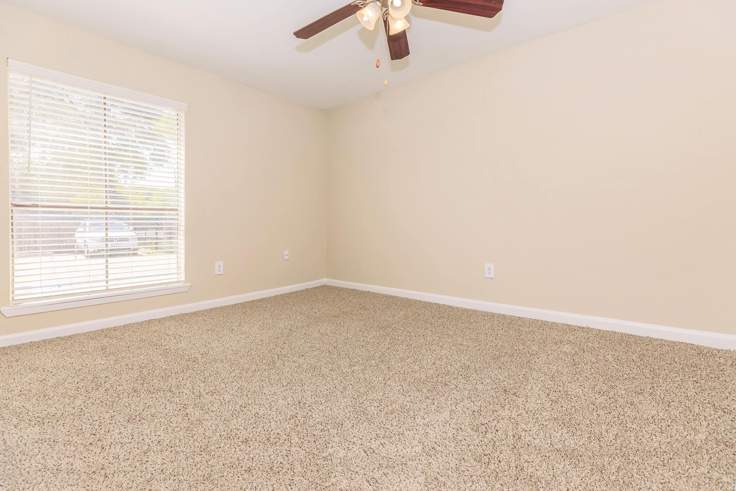 A bright, empty bedroom featuring beige walls, a light brown carpet, a ceiling fan with wooden blades, and a window with white blinds allowing natural light. The room is spacious, giving a clean and fresh feeling.