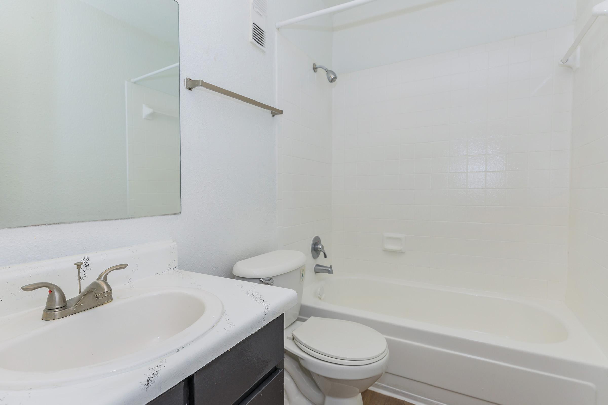 A clean bathroom featuring a bathtub and shower combination, a white sink with a faucet, and a toilet. The walls are light-colored, and there is a mirror above the sink. A towel bar is mounted on the wall, and the overall space appears bright and well-maintained.