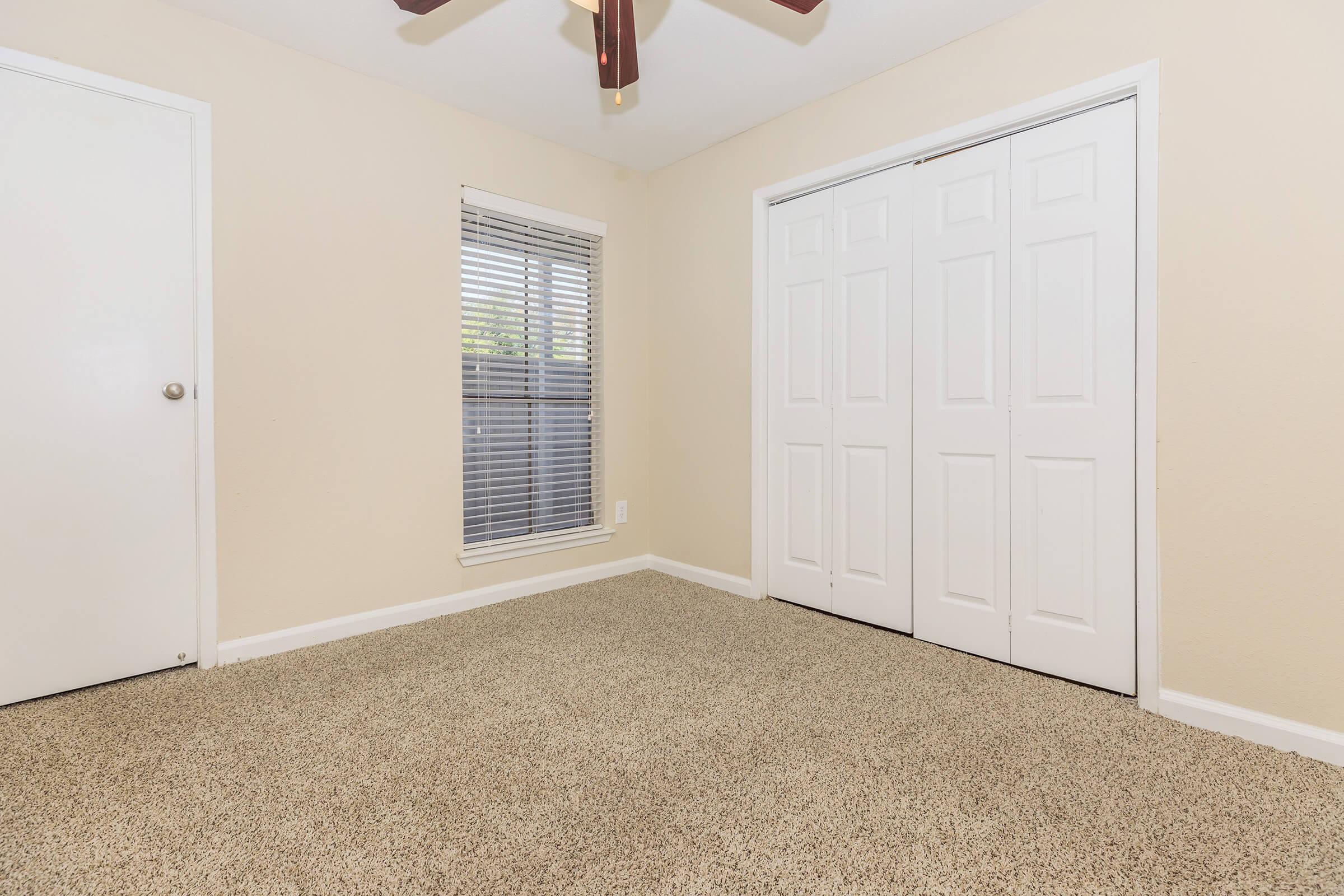 A simple, empty room with beige walls and carpet. There is a white door on the left, a window with blinds in the center, and a sliding closet door on the right. The ceiling features a brown ceiling fan, and the overall space appears clean and well-lit.