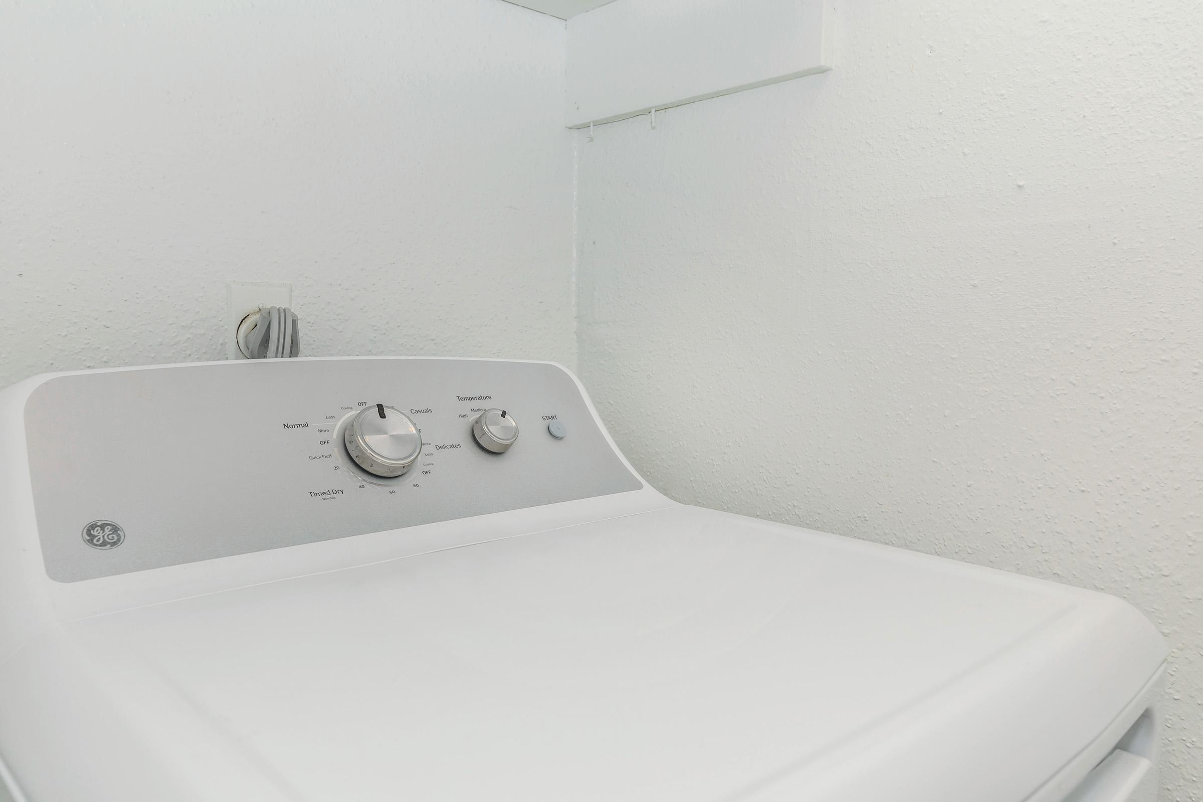 A white washing machine with control knobs sitting against a plain, lightly textured wall in a laundry area. The machine is empty and clean, showcasing its top surface and control panel, which indicate various settings for washing cycles.