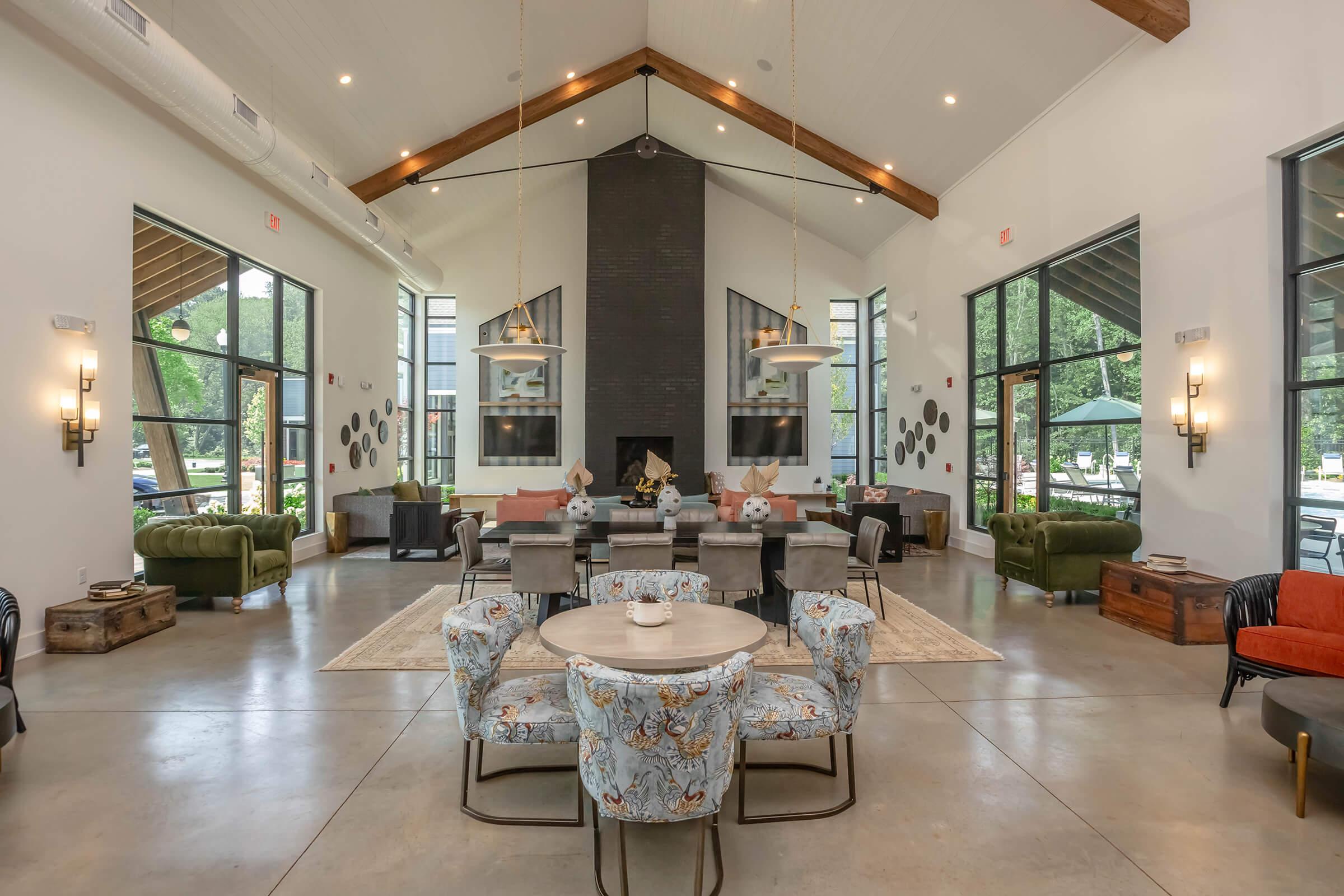 Spacious modern interior of a communal lounge area, featuring a high ceiling with wooden beams, stylish seating arrangements, a central fireplace, and large windows that let in natural light. Decorative elements and a cozy atmosphere create an inviting space for socializing and relaxation.
