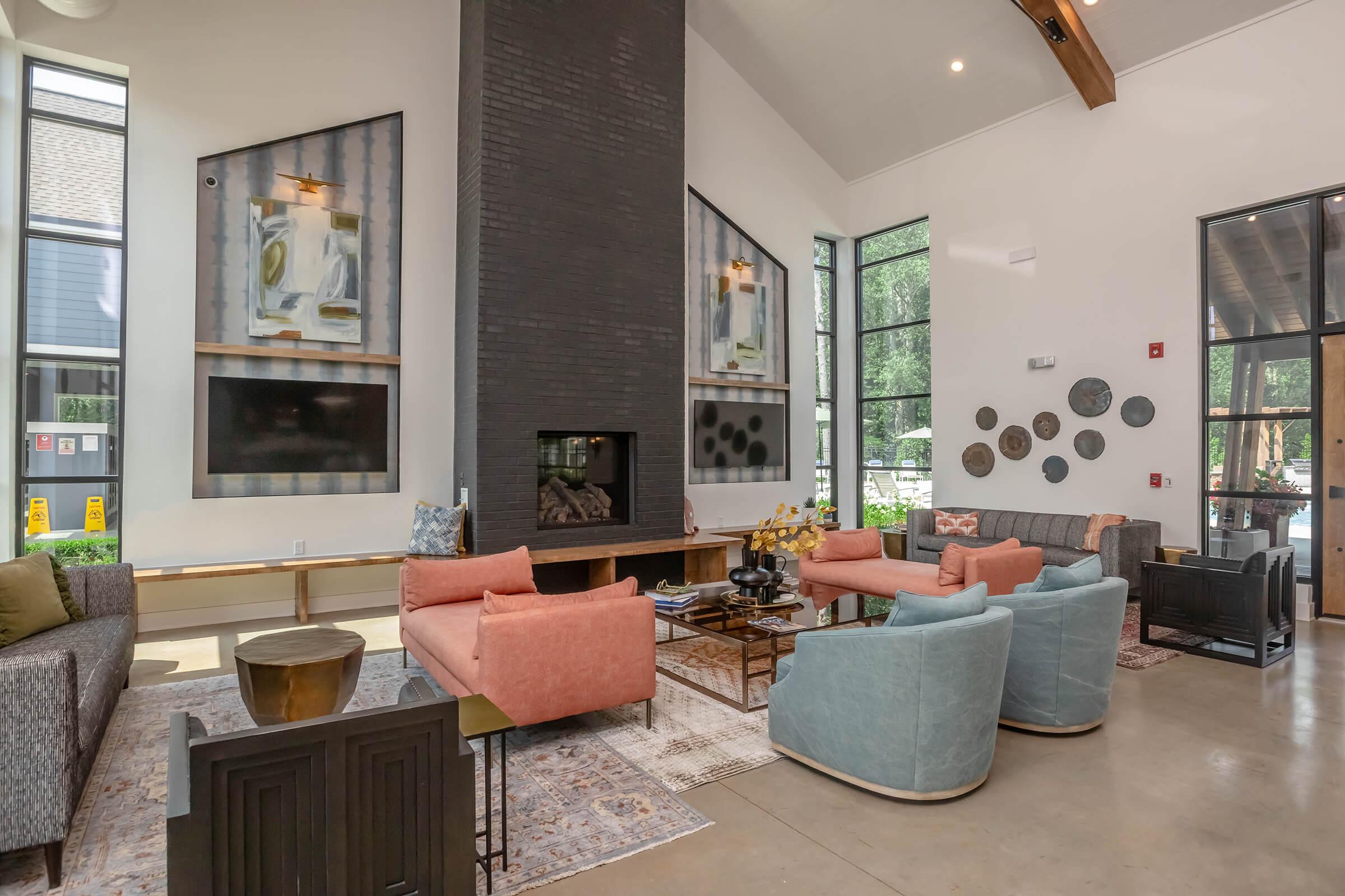A modern lounge area featuring colorful sofas in peach and blue, a central coffee table, and a fireplace embedded in a black feature wall. Large windows allow natural light, and artwork is displayed on the walls. The flooring is polished concrete, complemented by decorative pieces.