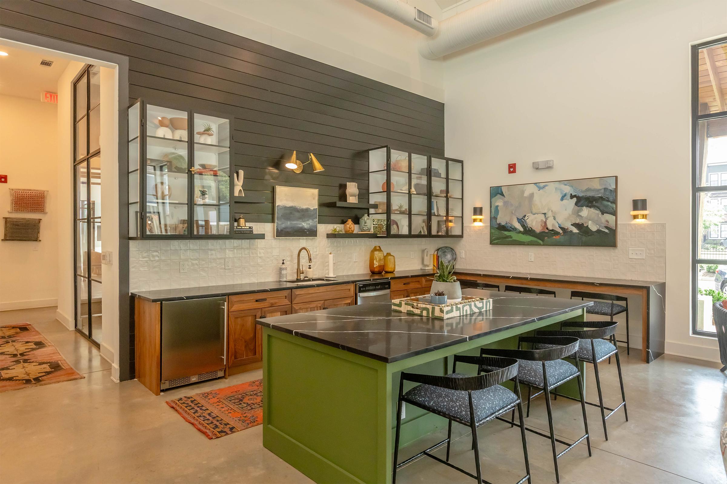 Modern kitchen with green island and black countertop, featuring glass-front cabinets displaying decor items. Wood cabinetry and light walls create a warm atmosphere, complemented by artwork on the walls. The space is well-lit with pendant lights and large windows. A patterned rug adds texture to the floor.