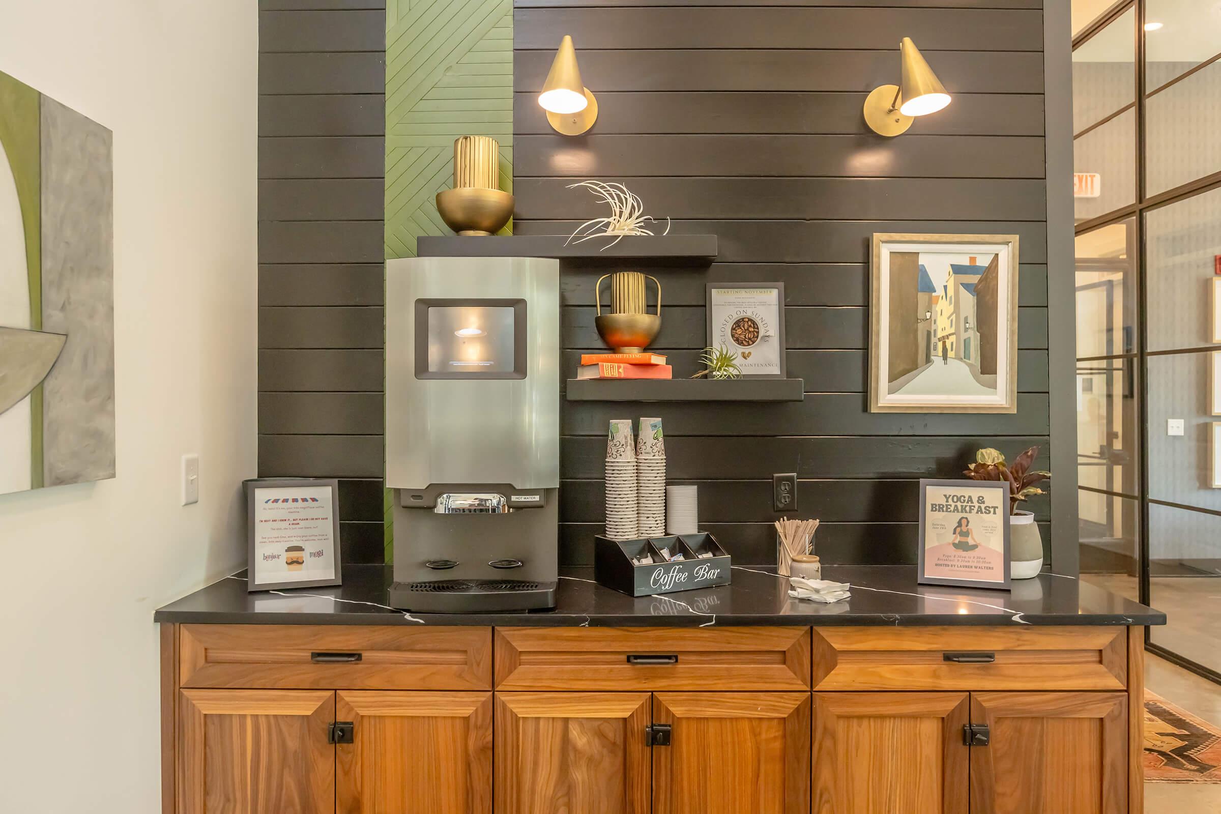Coffee station with a silver coffee machine, stacked paper cups, and a sign that reads "Coffee Bar." There are decorative elements including a plant, a framed picture of buildings, and two wall sconces providing ambient lighting. The background features a textured wall with green accents.
