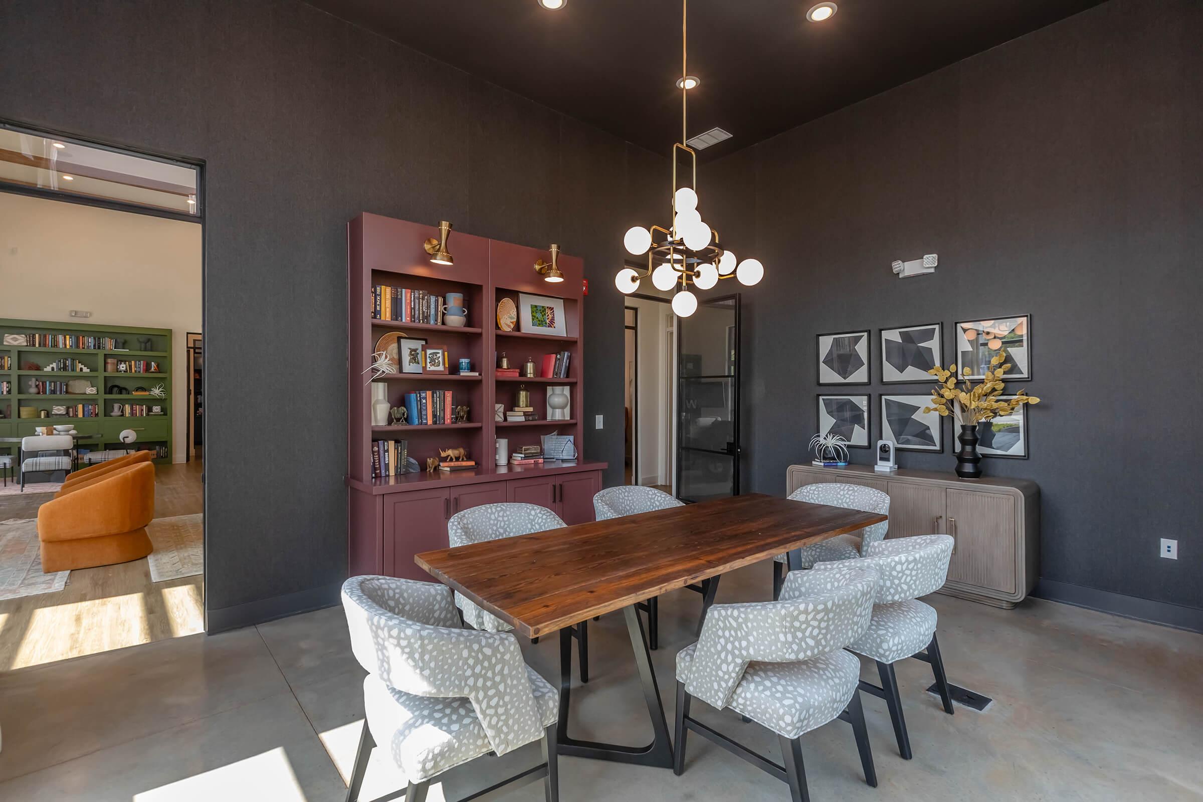 A modern dining area featuring a wooden table surrounded by six patterned chairs. A dark wall complements a stylish bookshelf filled with books and decorative items. A large pendant light hangs above the table, and framed artwork decorates the walls. Light enters from nearby windows, enhancing the inviting atmosphere.