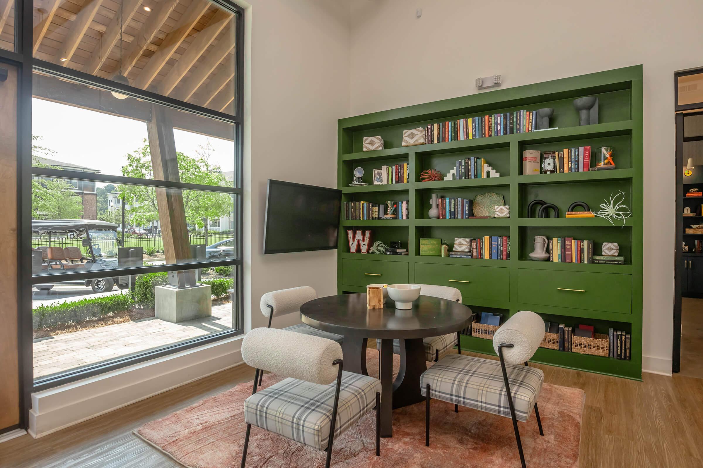 A stylish interior of a modern living space featuring a green bookshelf filled with books and decor items. In the foreground, a round black dining table surrounded by four upholstered chairs with a plaid pattern. Large windows allow natural light and offer views of the outdoors, creating a welcoming atmosphere.