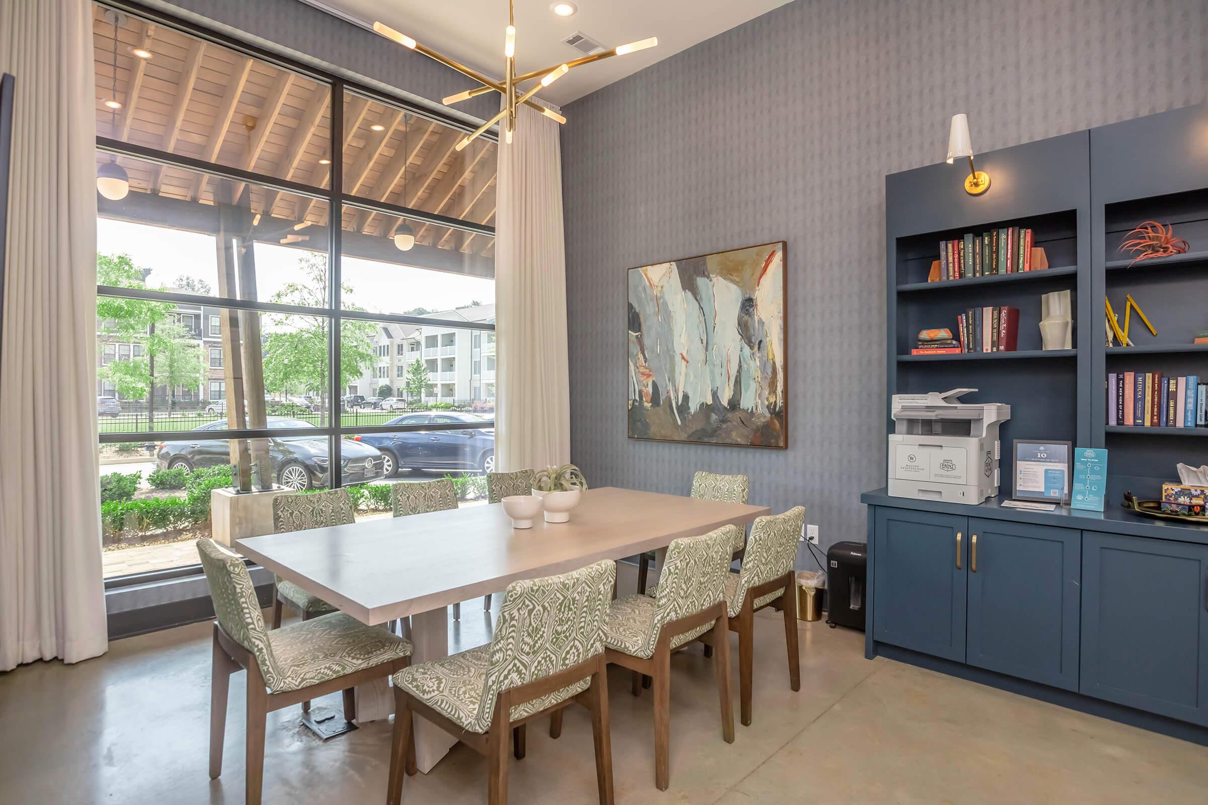 A modern dining area featuring a large wooden table surrounded by patterned chairs, illuminated by a stylish light fixture. In the background, large windows allow natural light to flood the space, revealing a view of greenery outside. A blue cabinet with books and decorative items adds a contemporary touch.