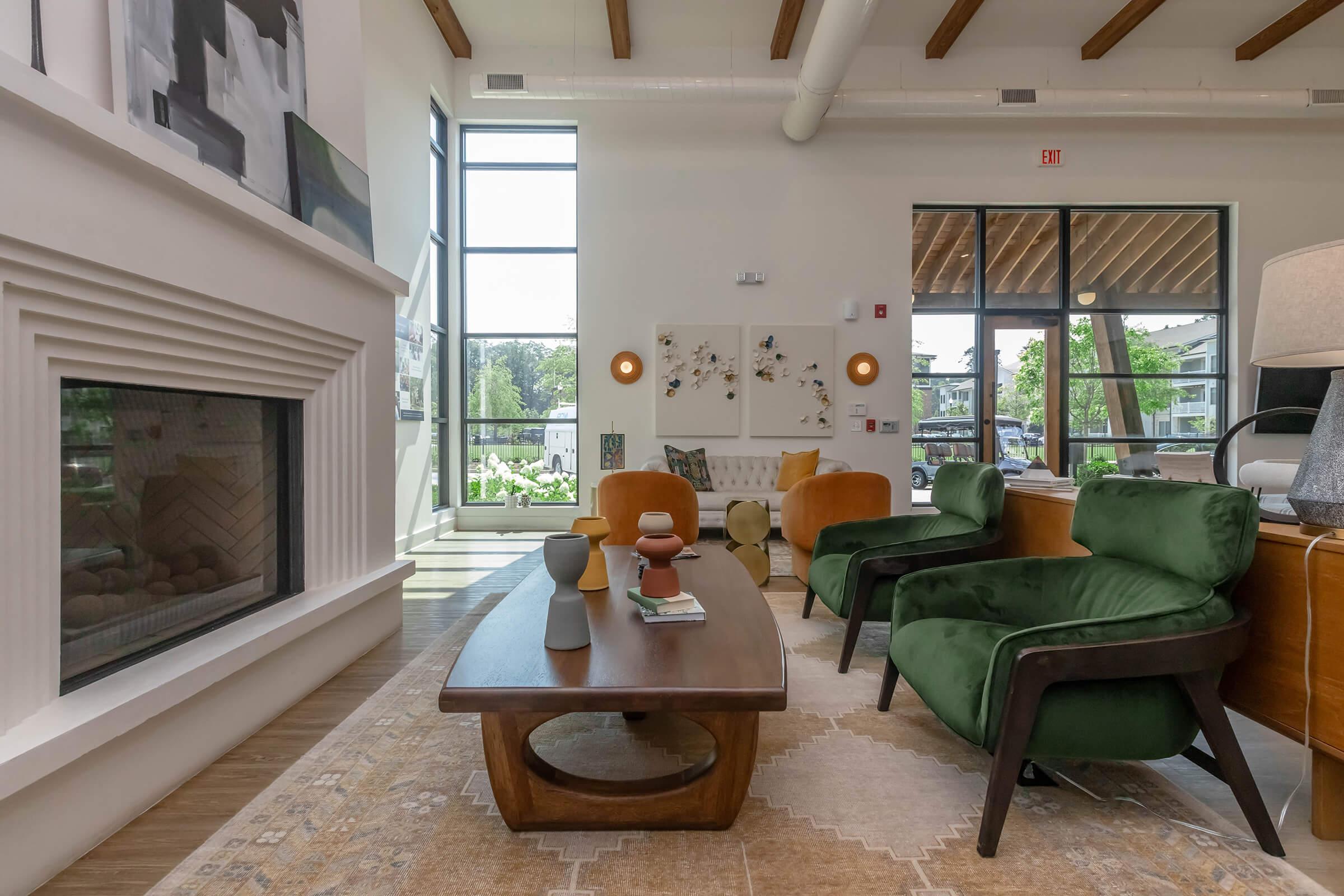 A modern living area featuring green armchairs and an orange sofa, with a wooden coffee table at the center. A fireplace is positioned against one wall, and large windows allow natural light to fill the space. Decorative artwork and plants enhance the cozy atmosphere.