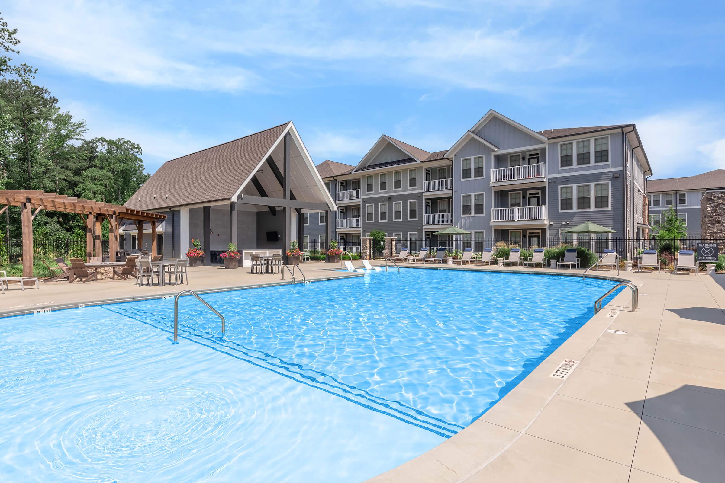A bright outdoor swimming pool with clear blue water, surrounded by lounge chairs and umbrellas. To the left, there’s a wooden pergola and seating area. In the background, a modern multi-story apartment building with balconies is visible, set against a clear blue sky and green trees.