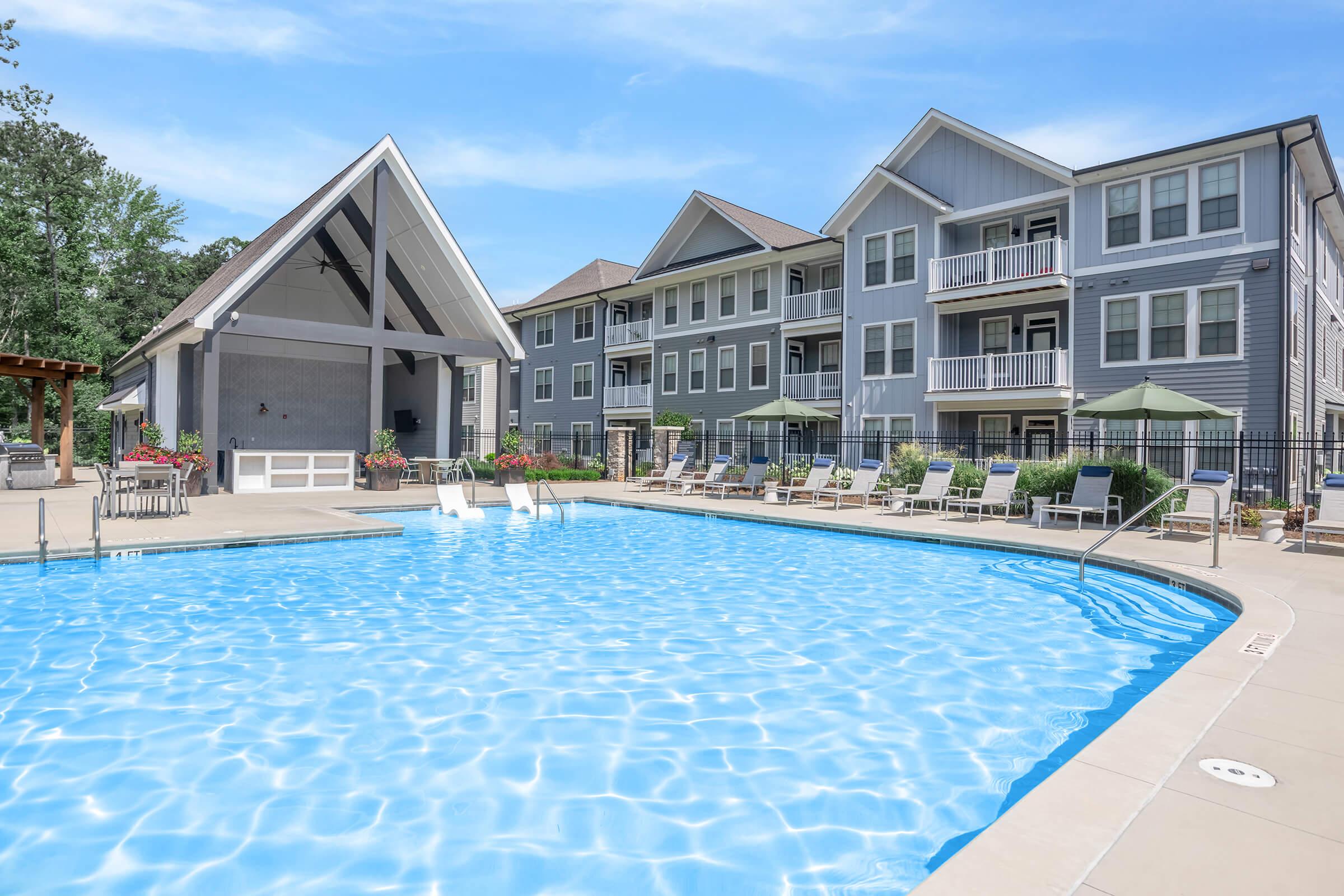A bright blue swimming pool surrounded by lounge chairs and umbrellas, with a large covered seating area and modern apartment buildings in the background. The scene is set in a landscaped outdoor area, featuring greenery and a clear blue sky.