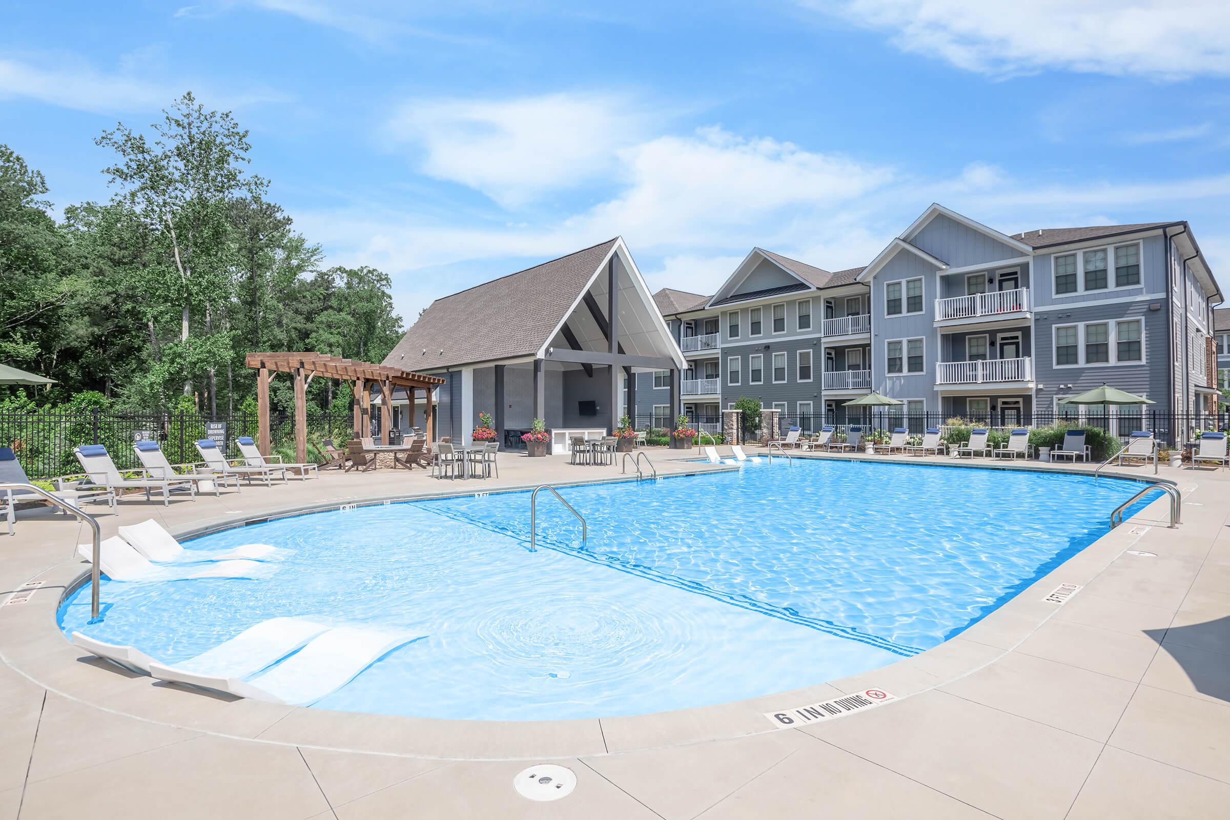 A spacious outdoor pool surrounded by lounge chairs and shaded areas. In the background, modern apartment buildings can be seen, along with well-maintained greenery. The sky is clear and sunny, creating a vibrant atmosphere ideal for relaxation and leisure.