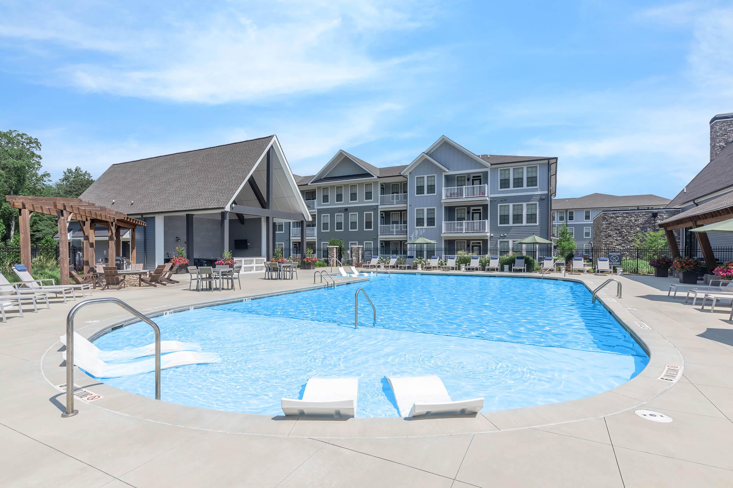 A sunny outdoor pool area surrounded by lounge chairs and shaded seating. The pool features a gentle curve, with steps leading in. Nearby, a modern building with multiple balconies and a charming pergola adds to the relaxing atmosphere in the background. Blue skies complete the scene.