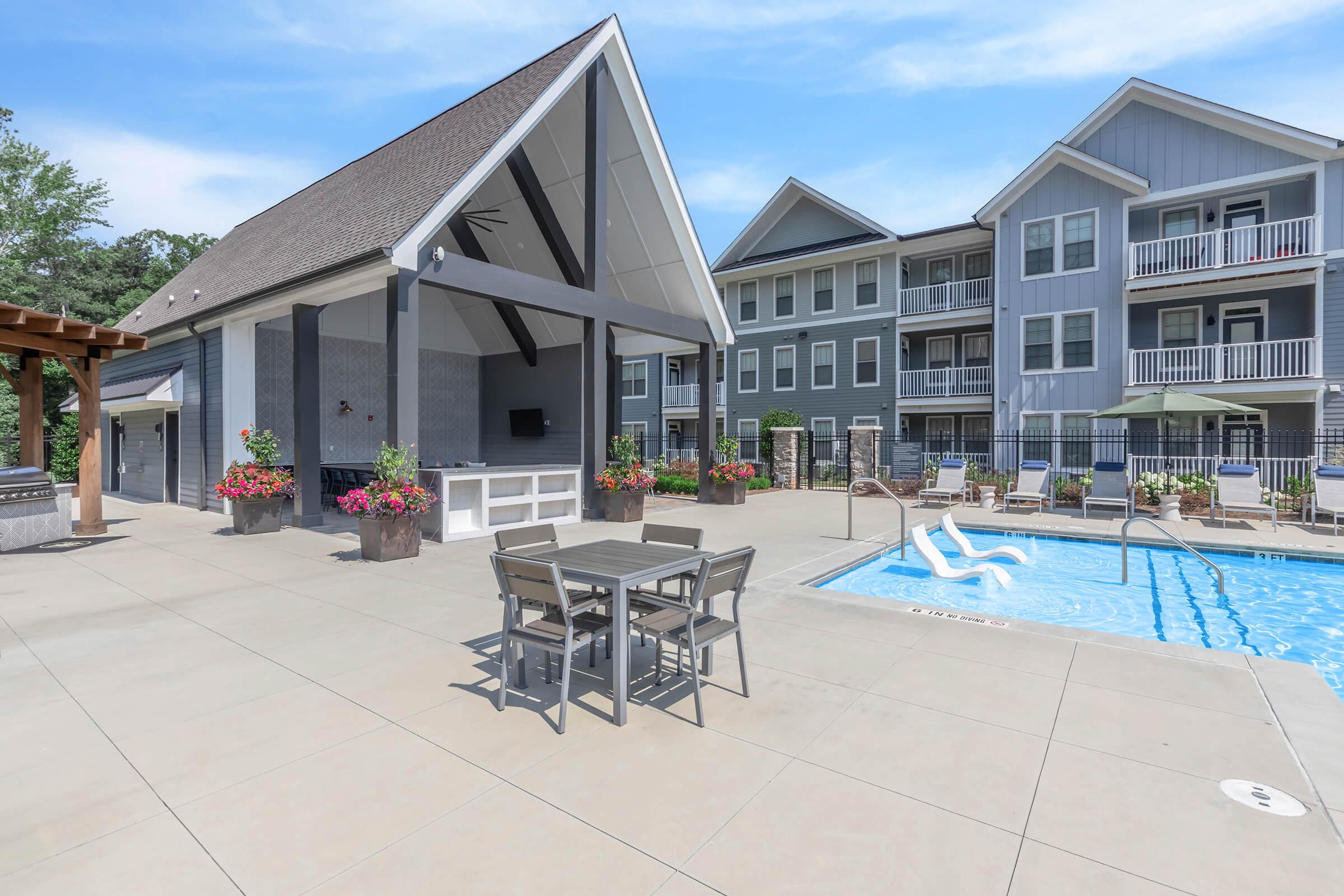 A modern outdoor pool area featuring a blue swimming pool, a seating area with metal tables and chairs, and a stylish pavilion. In the background, there are multi-story residential buildings with balconies. Lush greenery and colorful flower pots enhance the inviting atmosphere.