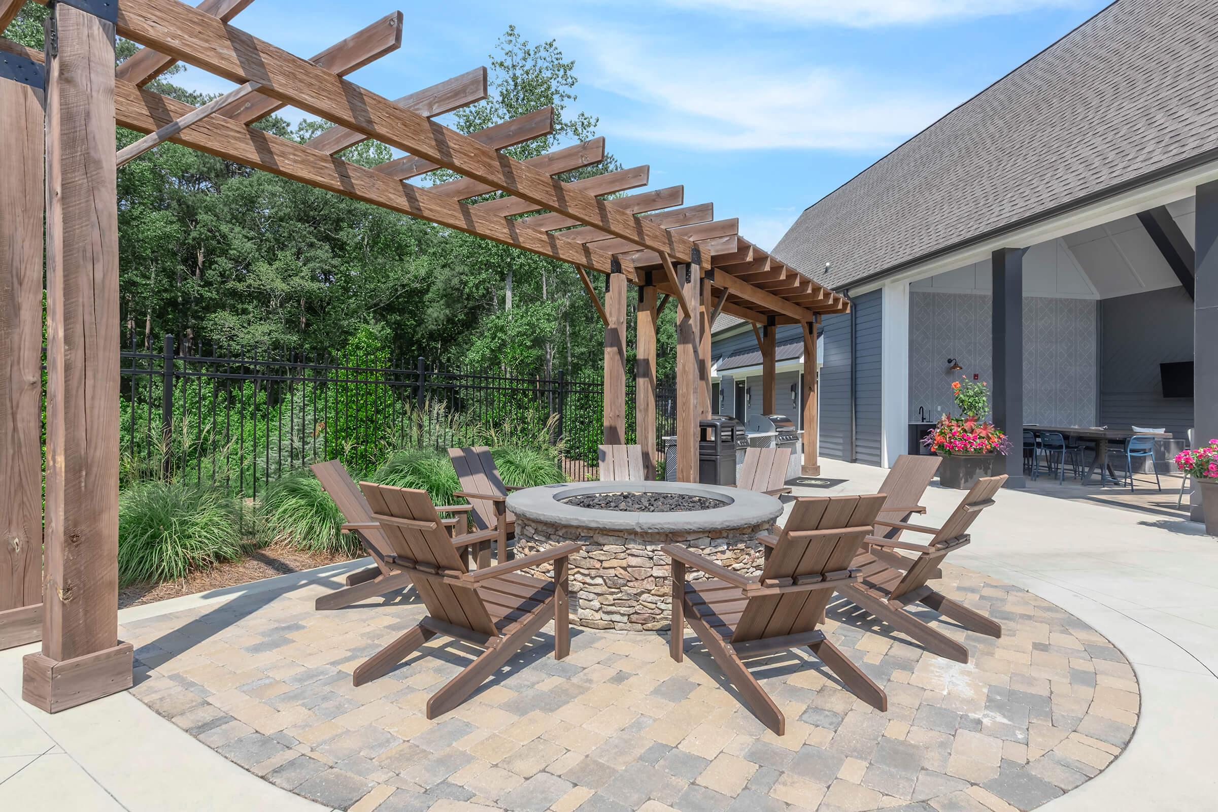 A cozy outdoor seating area featuring a circular stone fire pit surrounded by wooden chairs, under a wooden pergola. Lush greenery and flowers are visible in the background, with a modern building nearby, creating a welcoming and tranquil atmosphere.