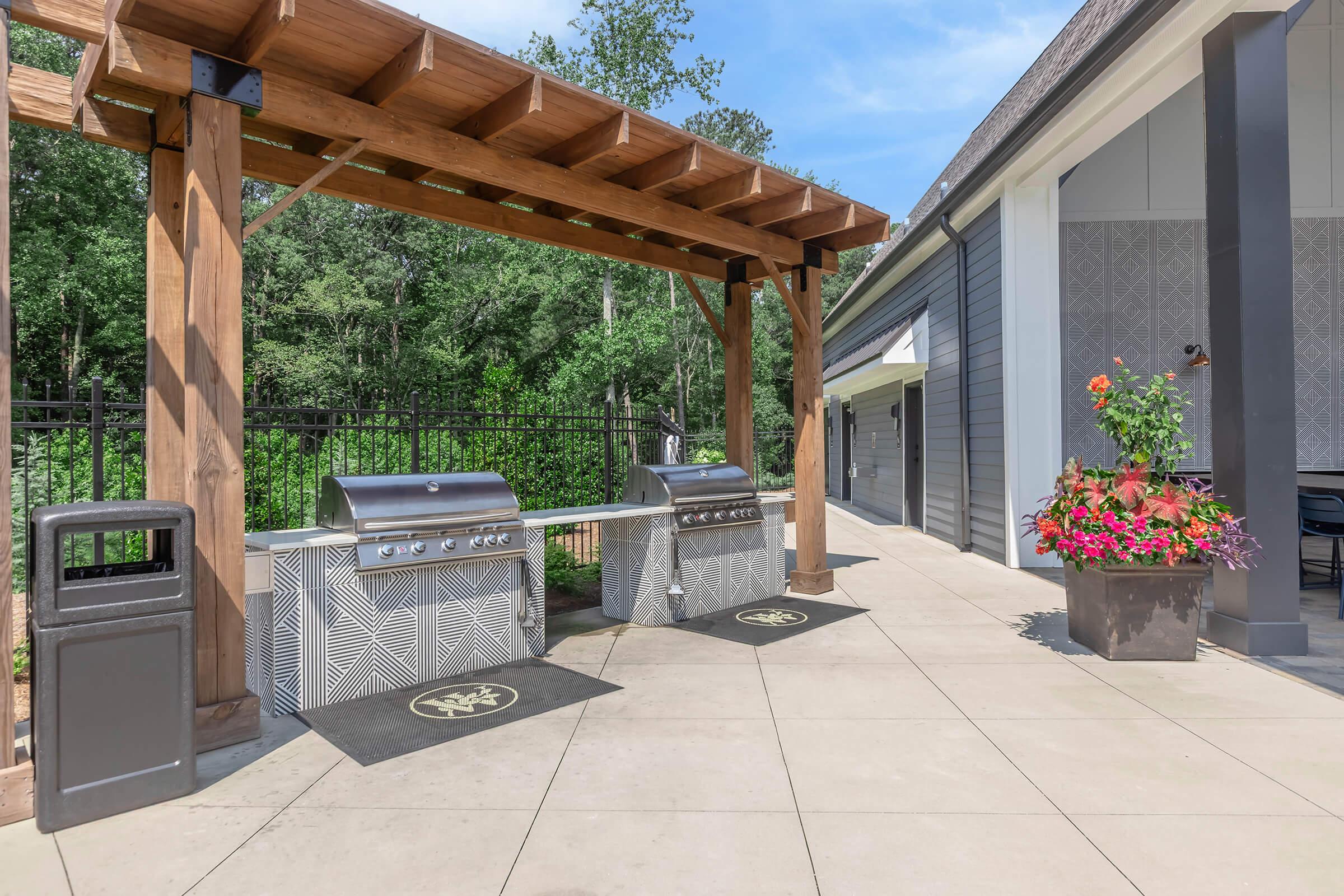 Outdoor grilling area featuring two stainless steel BBQ grills under a wooden pergola. The space includes a shaded patio with tiled flooring, a decorative planter filled with colorful flowers, and a trash can nearby. Lush greenery is visible in the background, creating a serene environment.