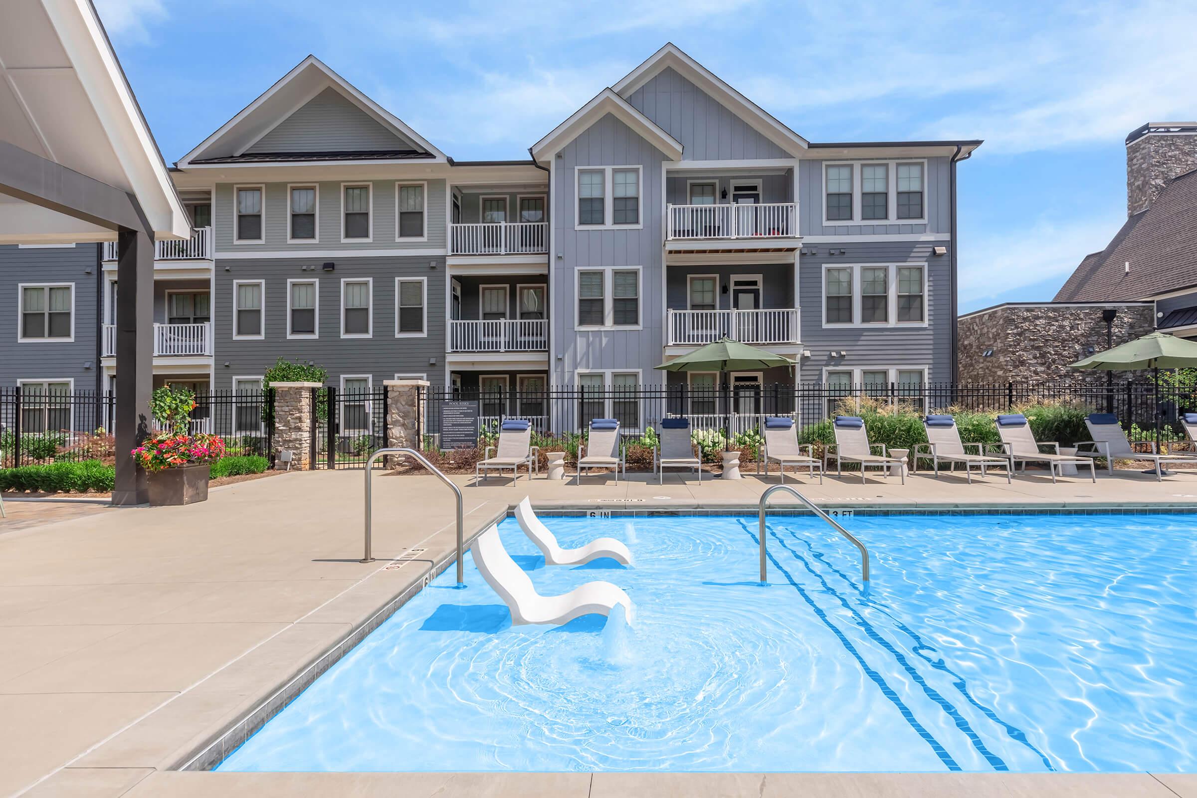 A sparkling blue swimming pool with chaise lounge chairs around it, situated in front of a multi-story apartment building. The area is well landscaped with potted plants and umbrellas, offering a relaxing atmosphere on a sunny day. The architecture is modern and inviting.