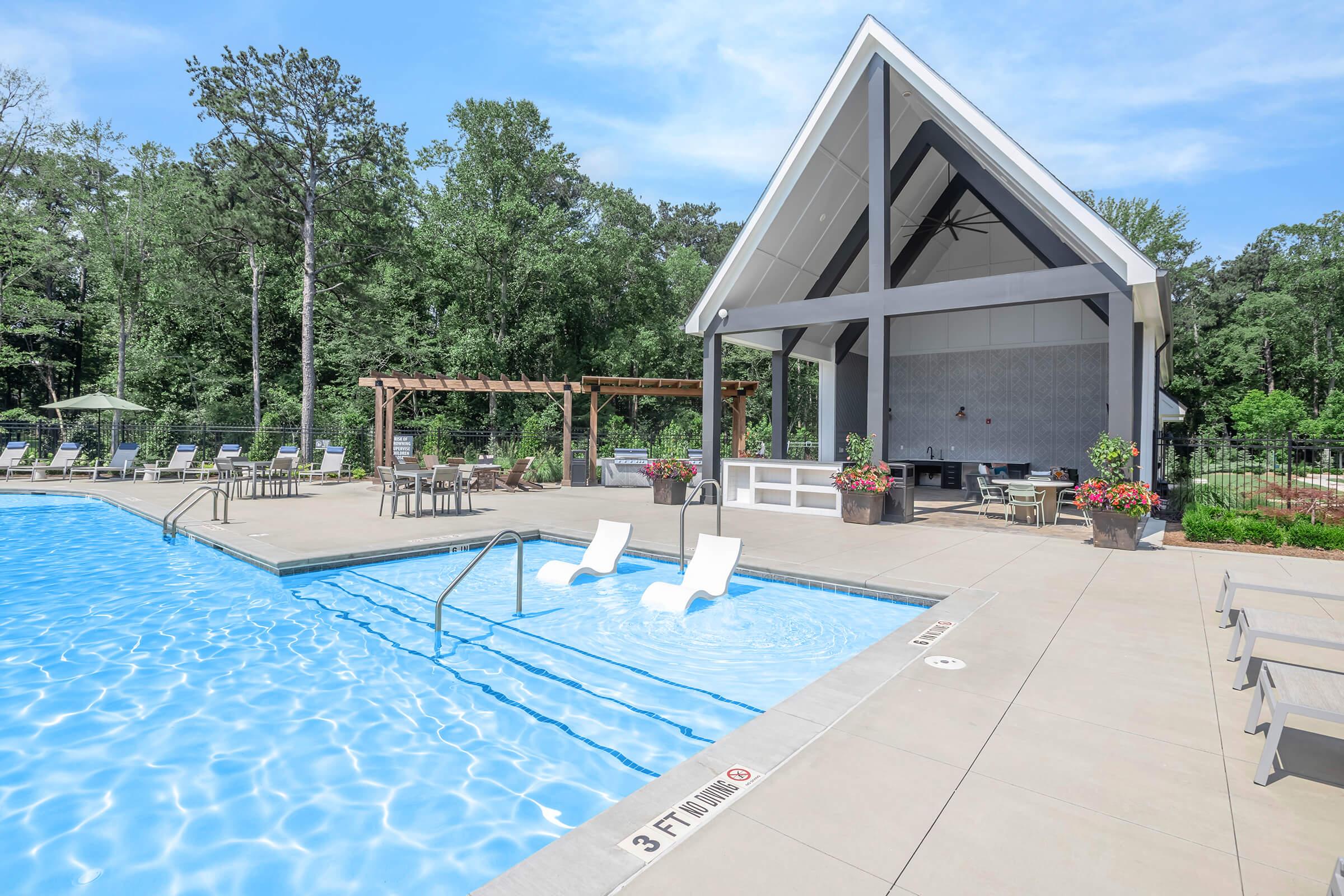 A modern pool area with clear blue water, featuring lounge chairs and a shaded seating area. There's a wooden pergola in the background surrounded by trees. The pool has clear depth markings, and flowers in planters add color to the scene.